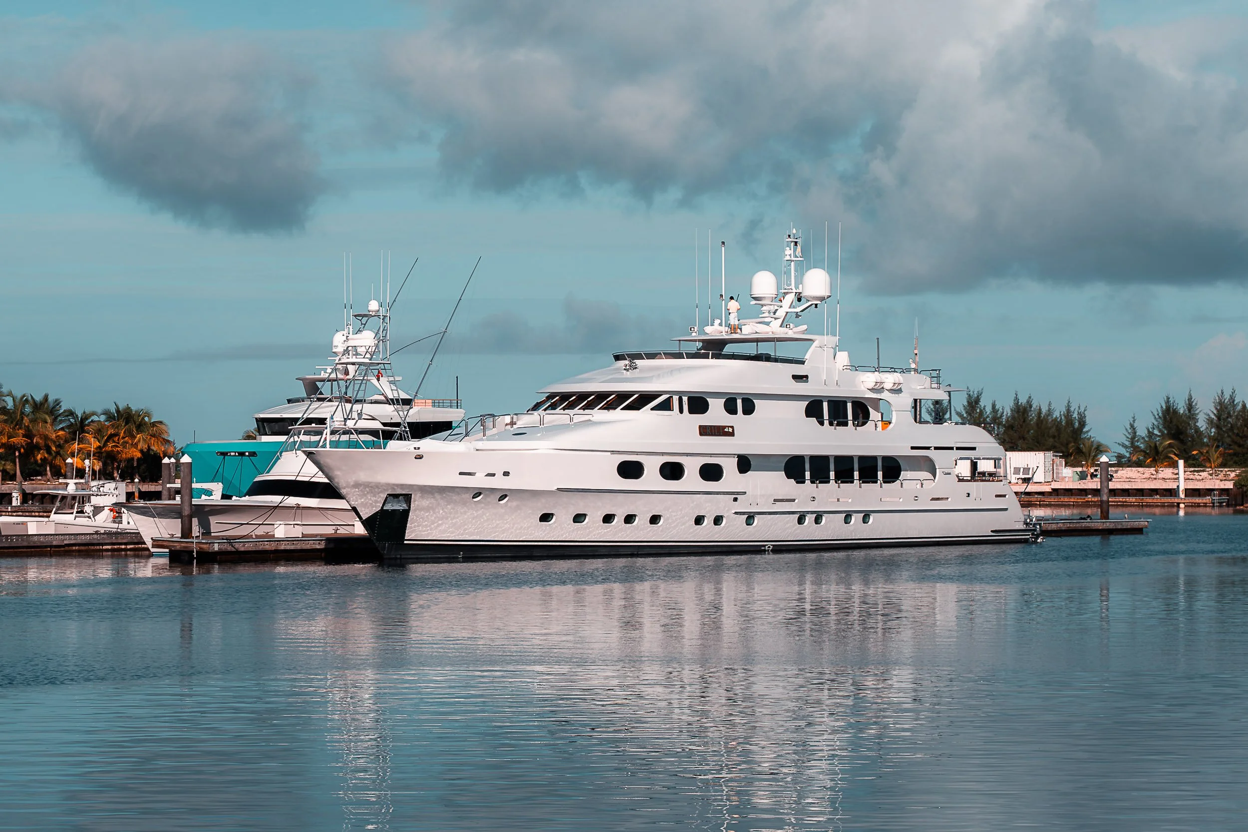 A large white yacht docked at a marina with smaller boats and palm trees in the background, under a partly cloudy sky.