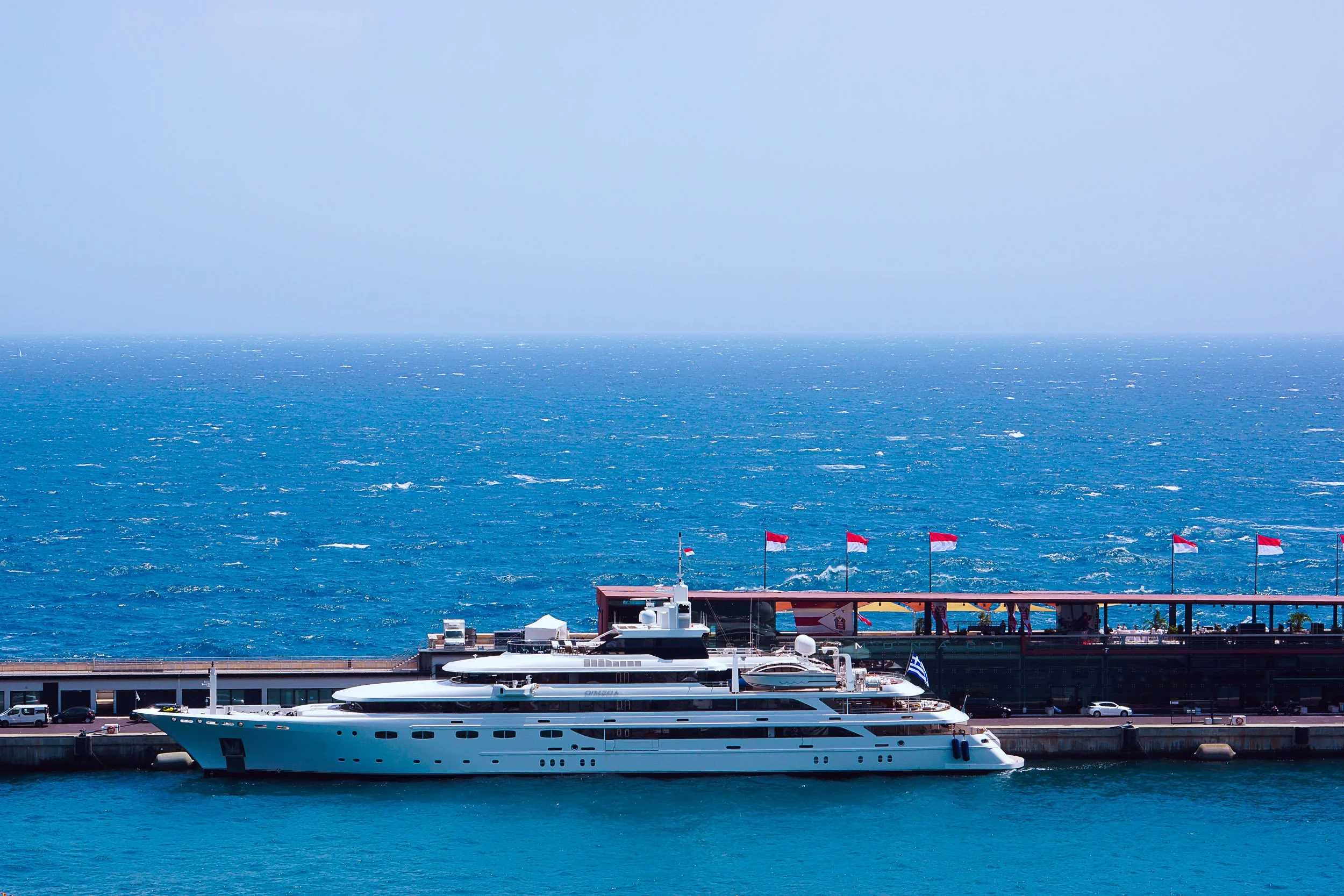 Luxury yacht docked at a pier with flags, set against a vast ocean backdrop.