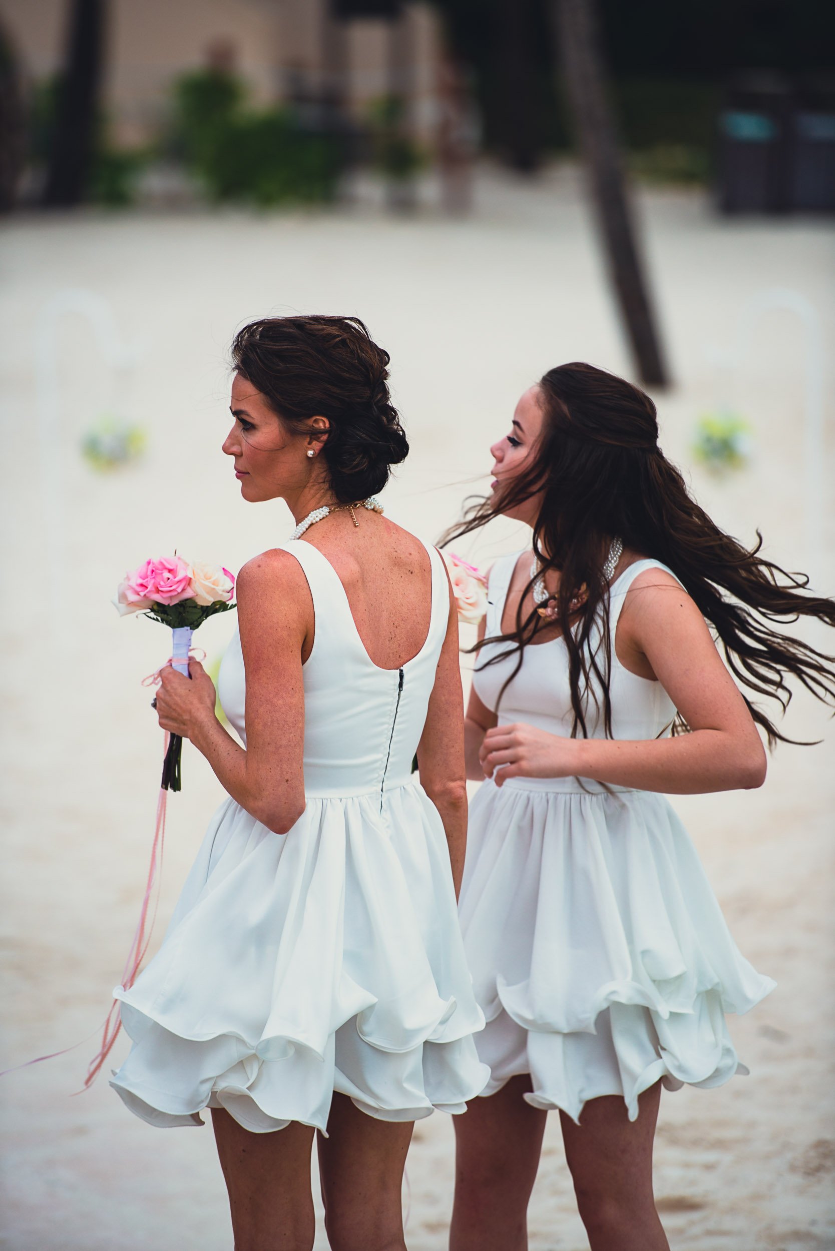 Two women in white dresses at a beach wedding holding flowers.