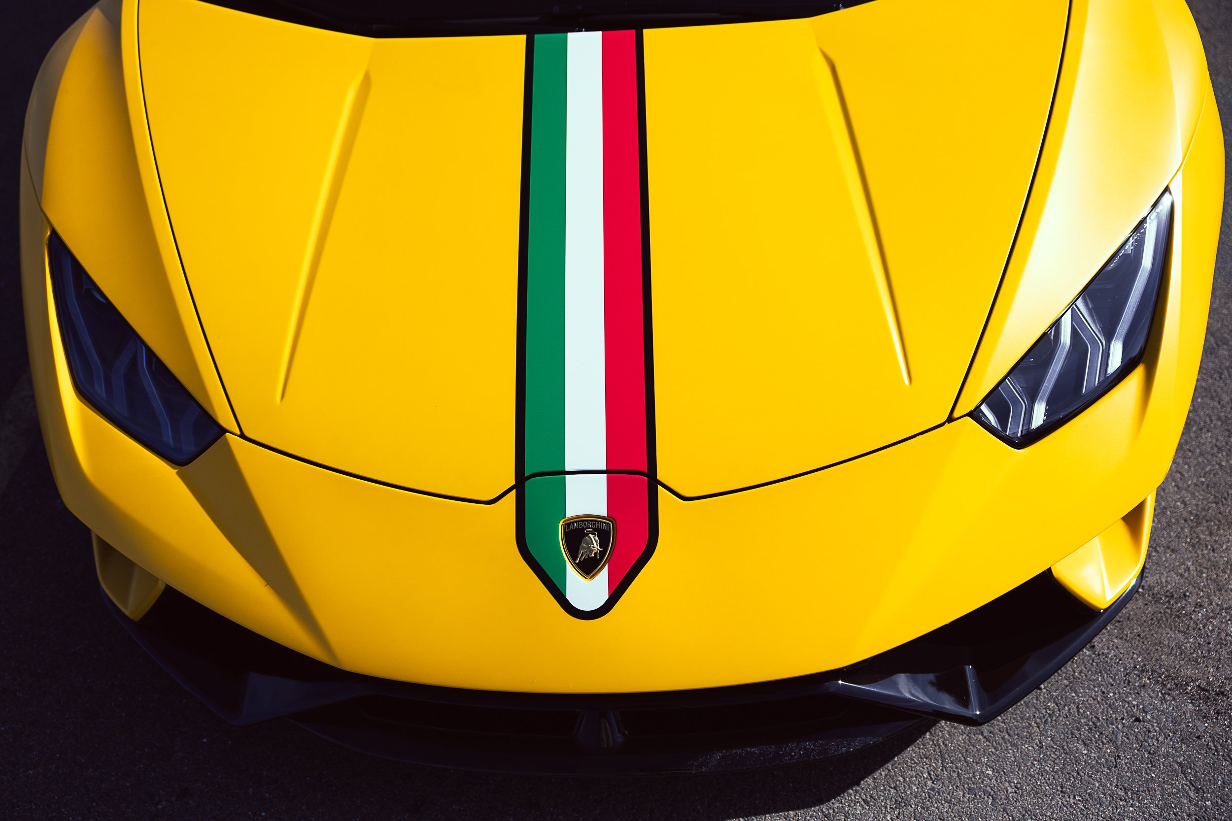 Close-up of the front of a bright yellow Lamborghini sports car with black accents, featuring a black and yellow Lamborghini emblem and an Italian flag stripe down the center.