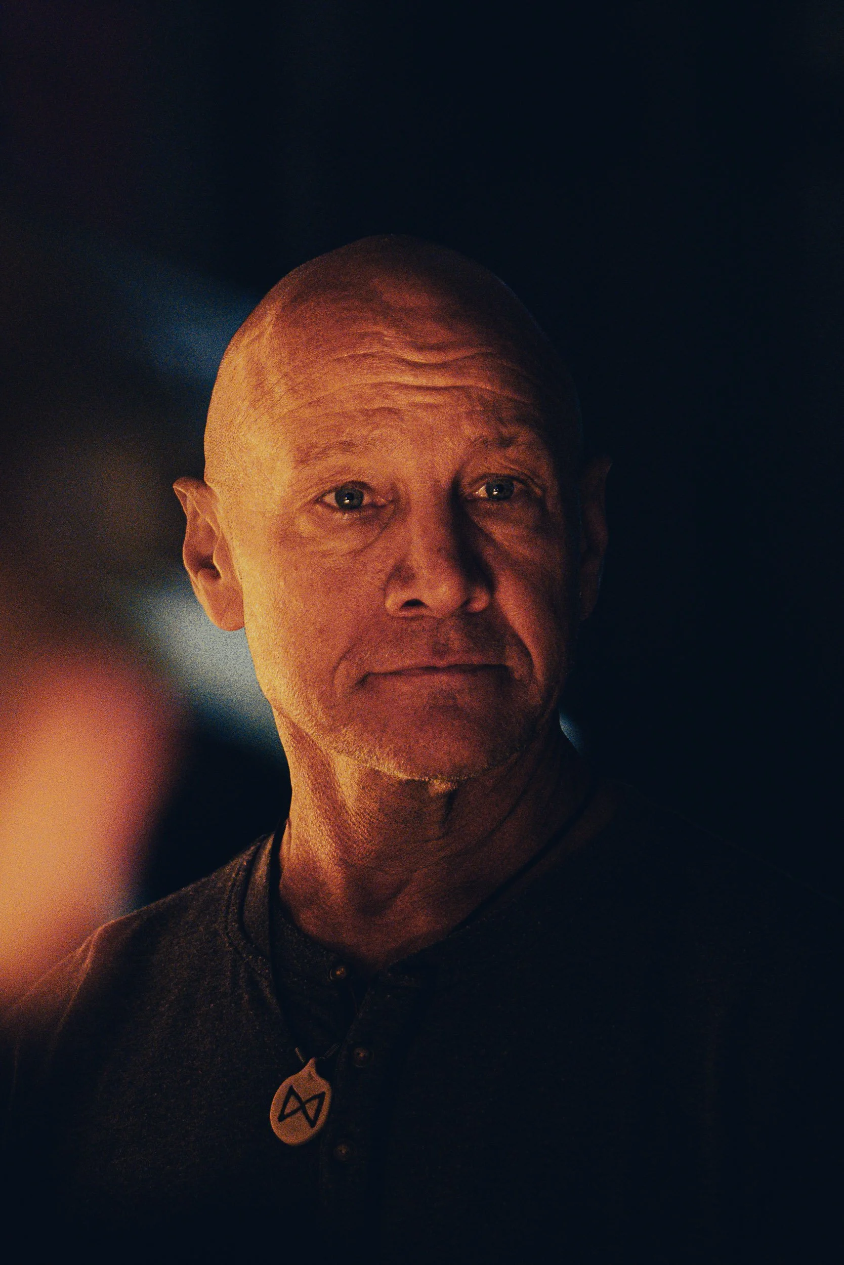 Close-up portrait of a bald man in dim lighting wearing a necklace.