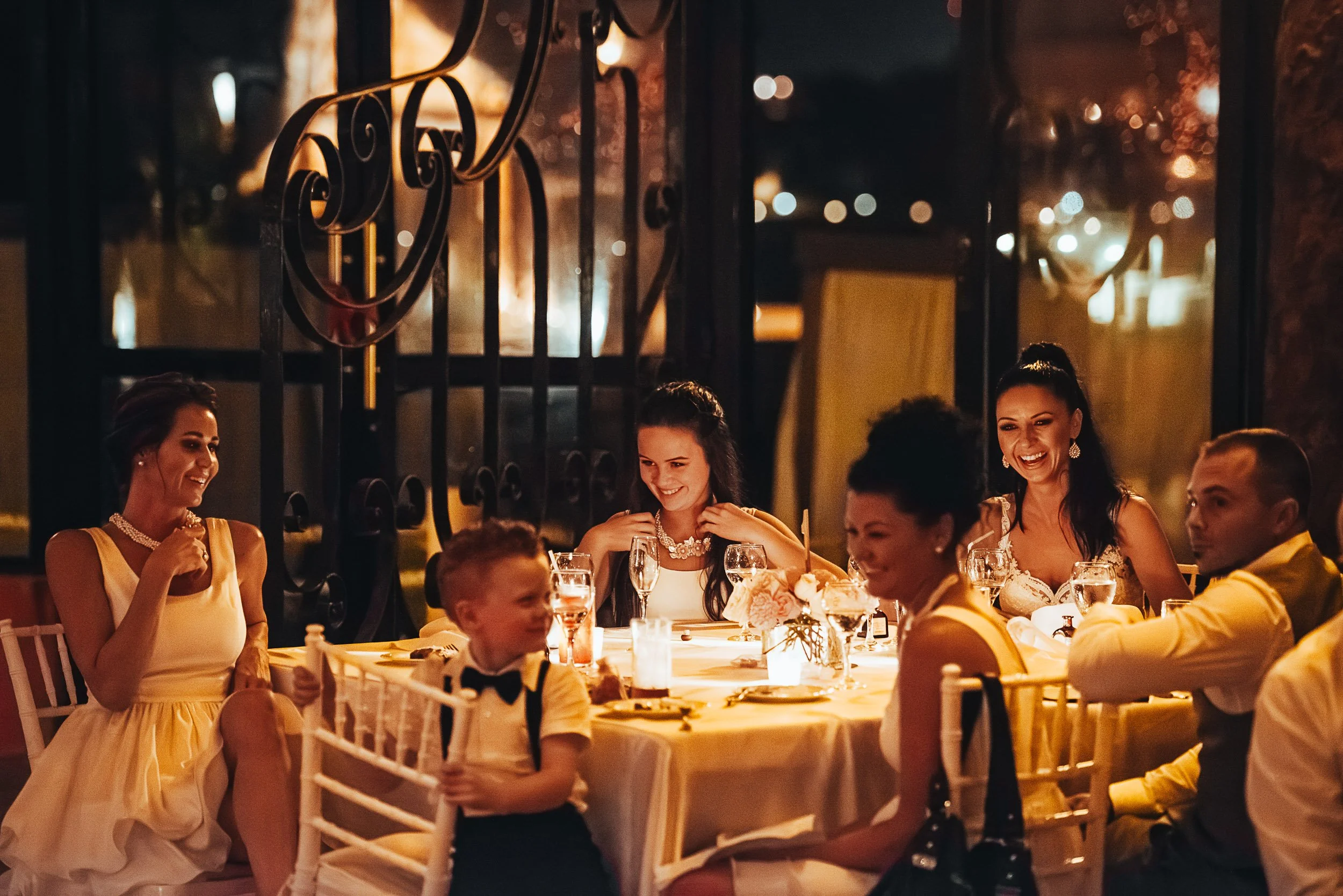 A group of people sitting around a table at a formal event, smiling and laughing in a warmly lit venue. The table is elegantly set with candles and flowers.