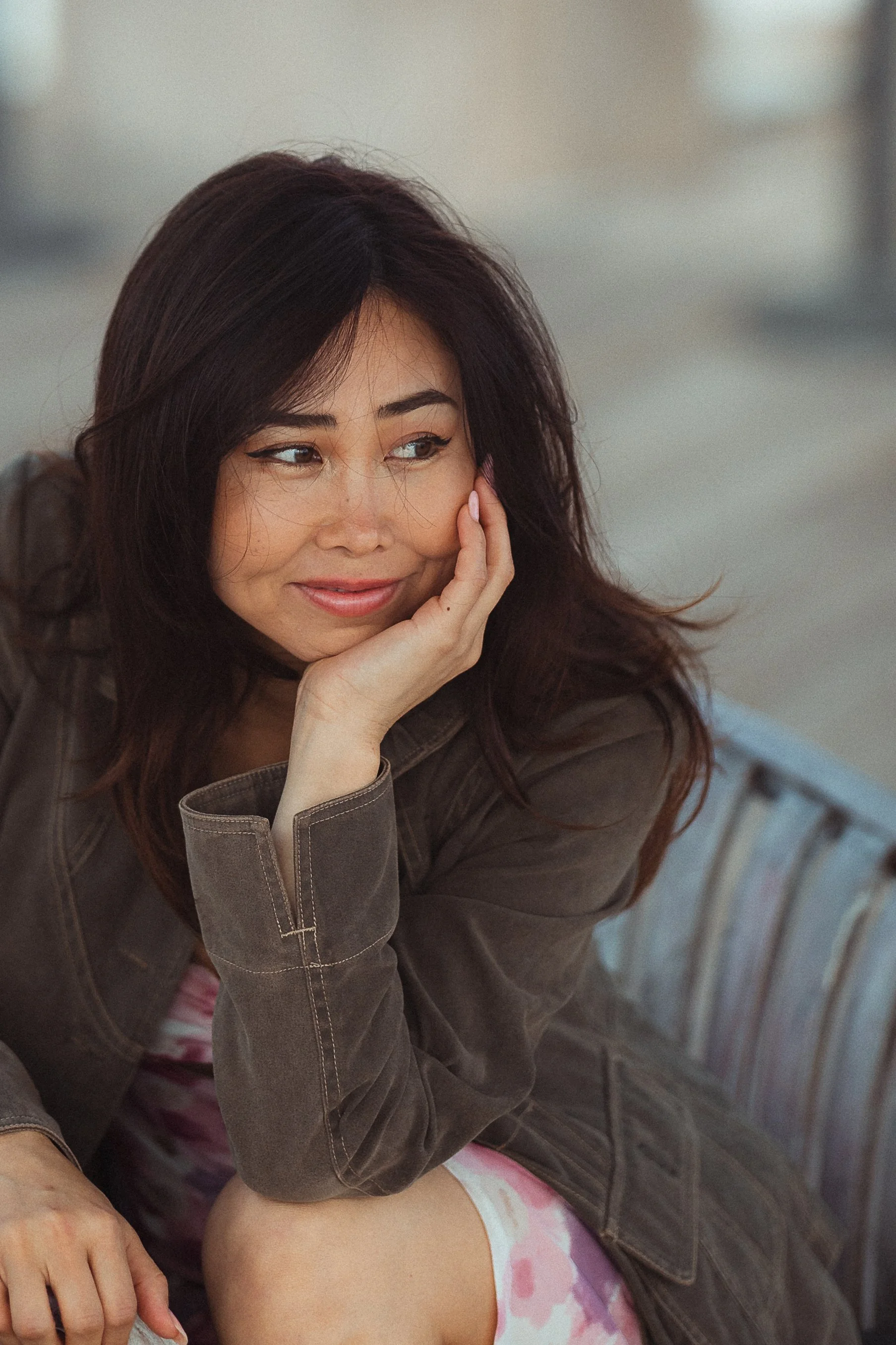 A young woman with dark brown hair and light skin is resting her face on her hand, smiling softly. She is wearing a brown jacket and a pink tie-dye shirt, sitting on a striped couch with blurred background.