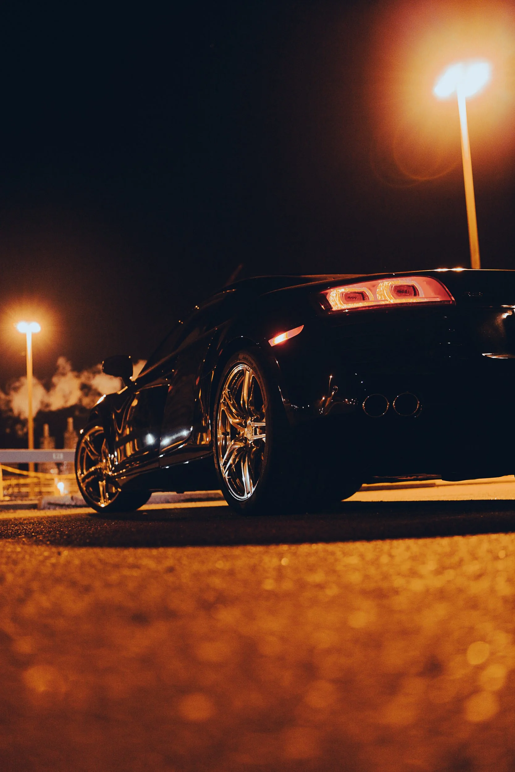 Low-angle view of a black sports car with illuminated tail lights parked under streetlights at night.