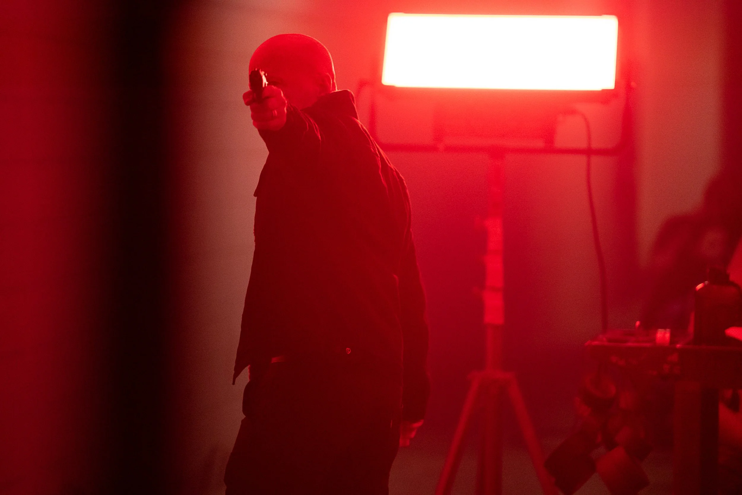 Person holding a gun in a dimly lit room with red lighting, standing near a bright light source on a tripod.