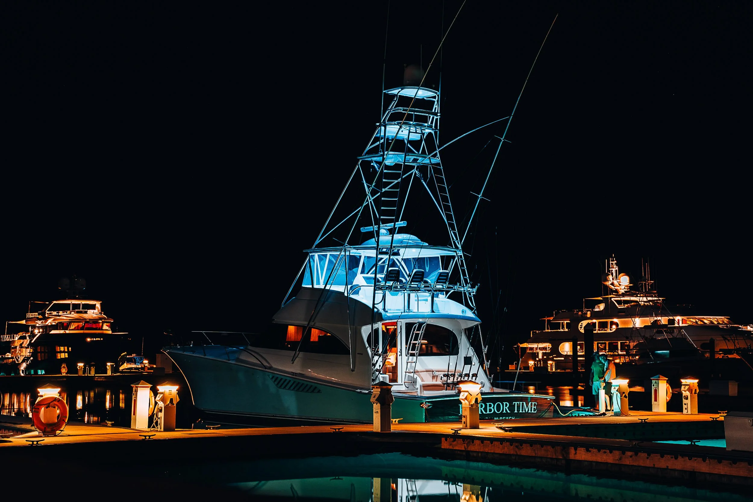 A marina at night with a luxury yacht and other boats docked, illuminated by bright lights. A person stands on the dock near the yacht.