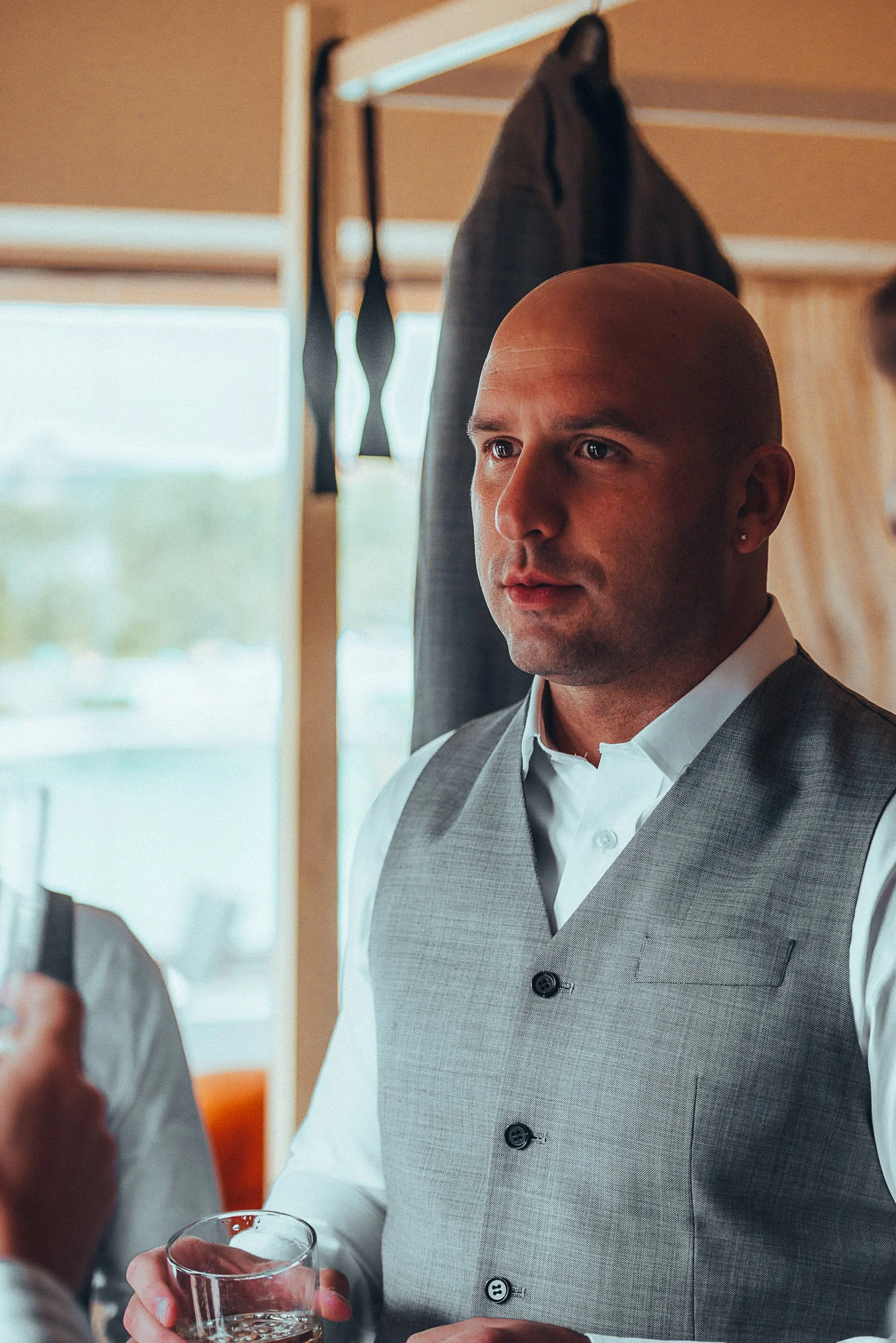 Man in gray vest holding a drink, with a suit jacket hanging in the background.