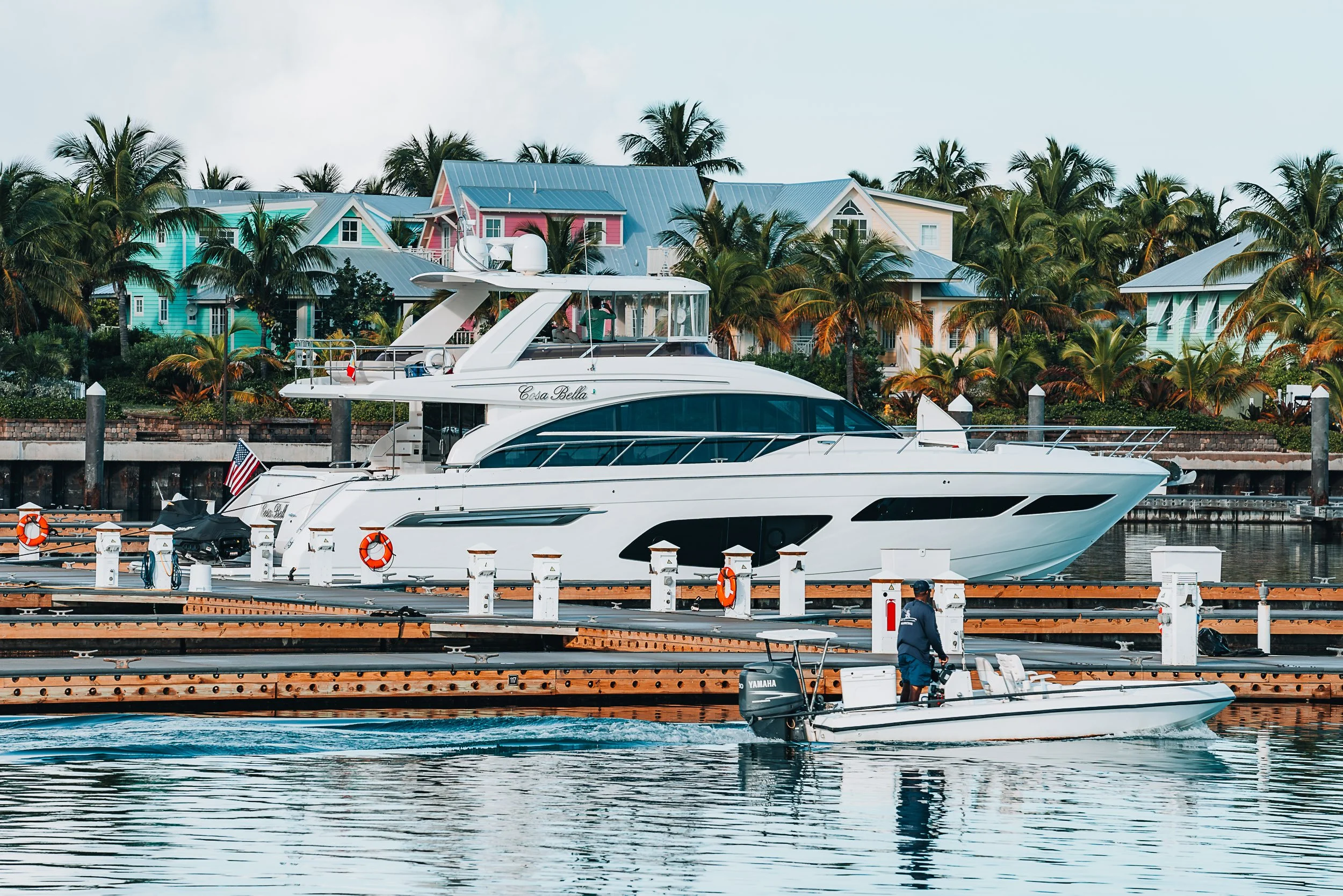 Luxury yacht docked at marina with colorful houses and palm trees in the background.