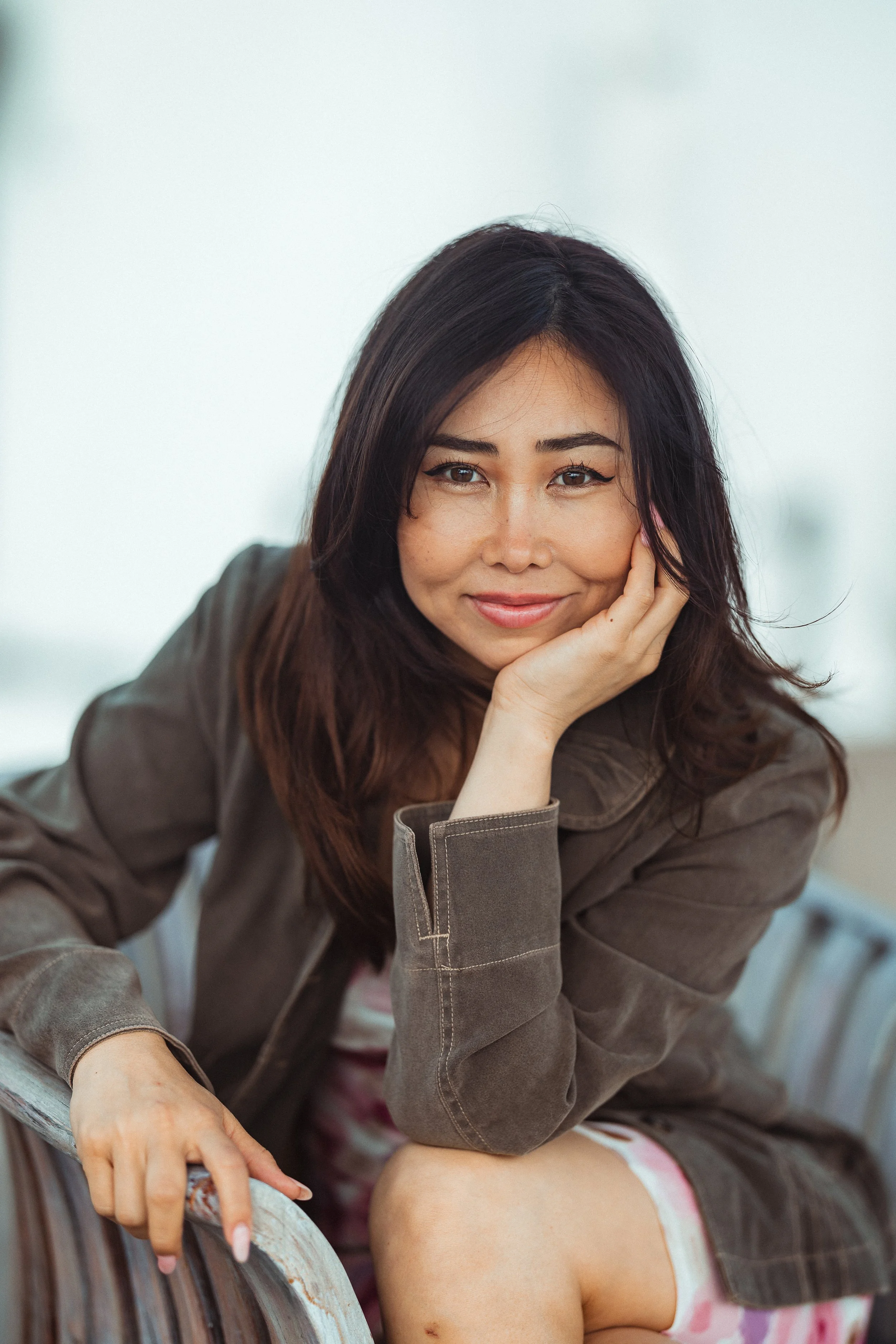 A woman with dark hair and a smile resting her chin on her hand, sitting outdoors on a bench with a child in a pink and white tie-dye shirt nearby.