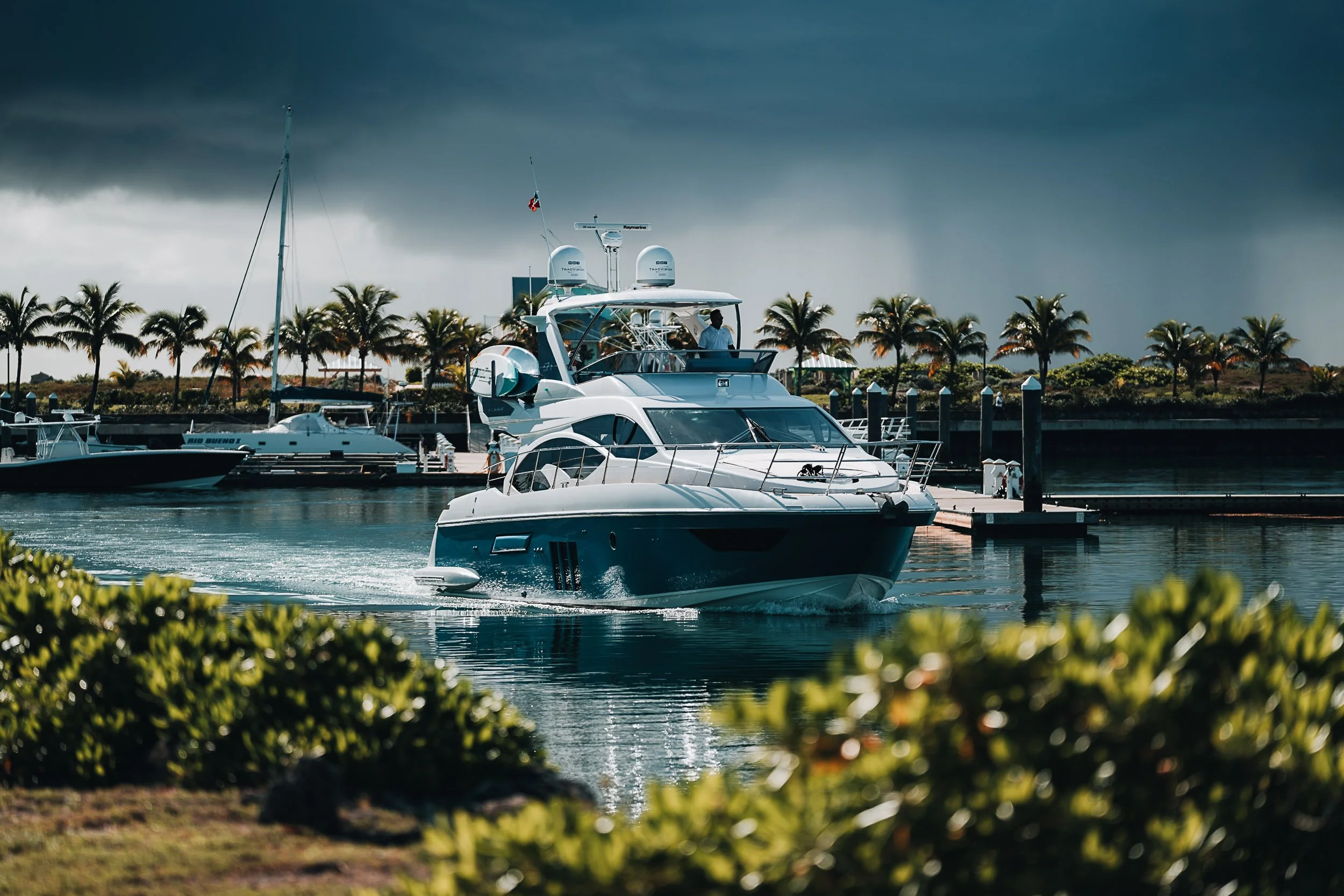 Luxury yacht sailing in marina with palm trees and dark clouds.