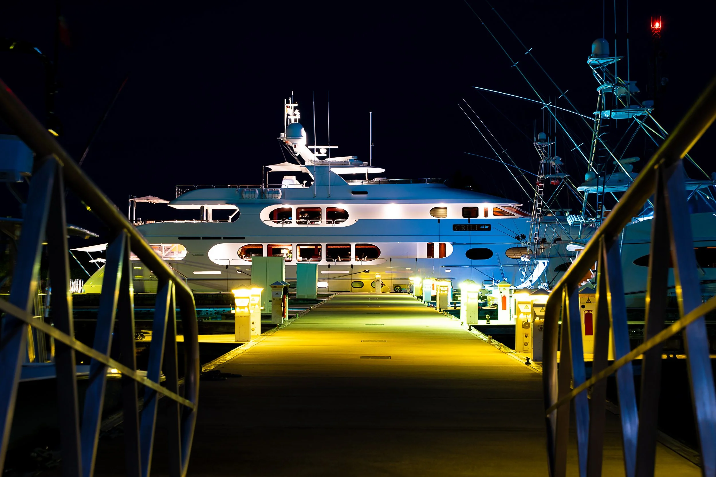 Luxury yacht docked at a marina at night, illuminated by dock lights, with rigging visible against a dark sky.