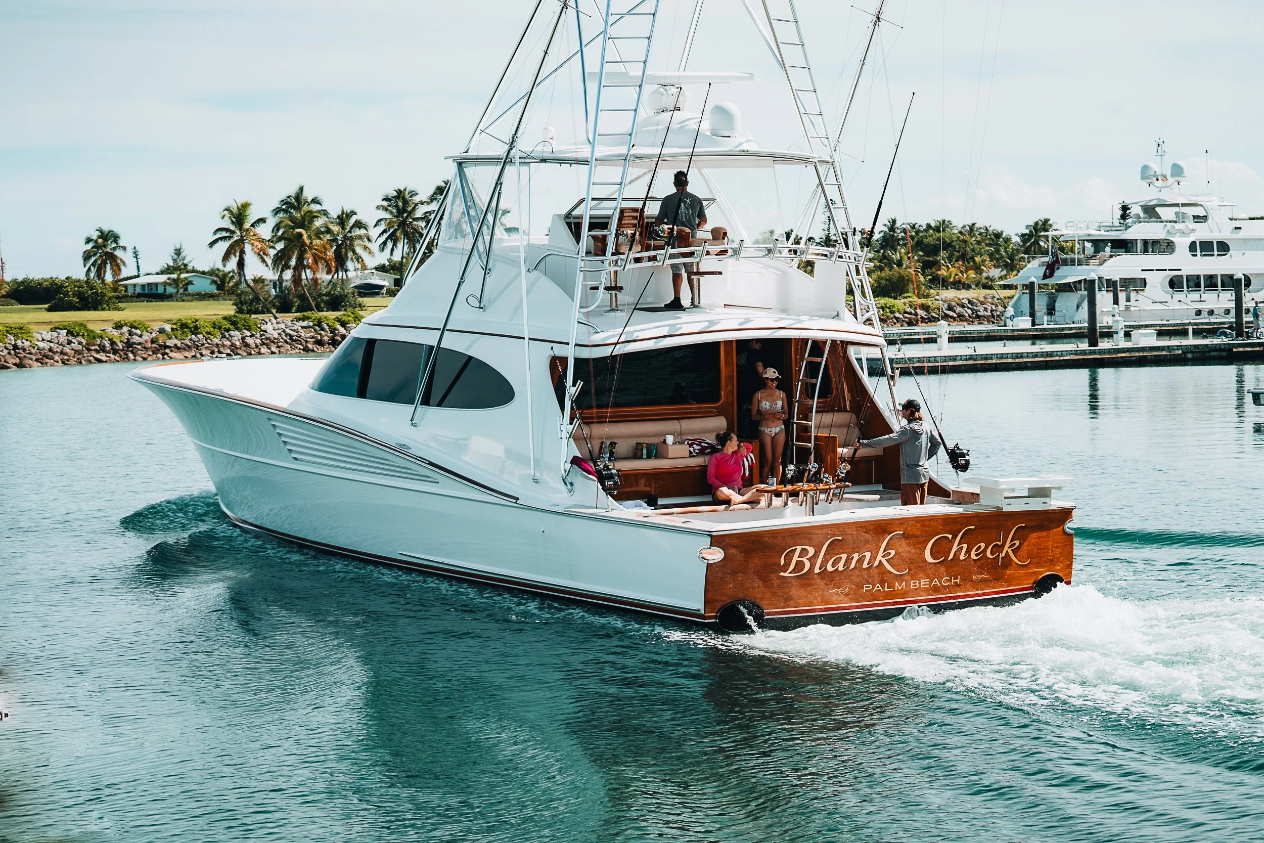 Luxury yacht with people onboard, cruising near a harbor with palm trees and other boats in the background.