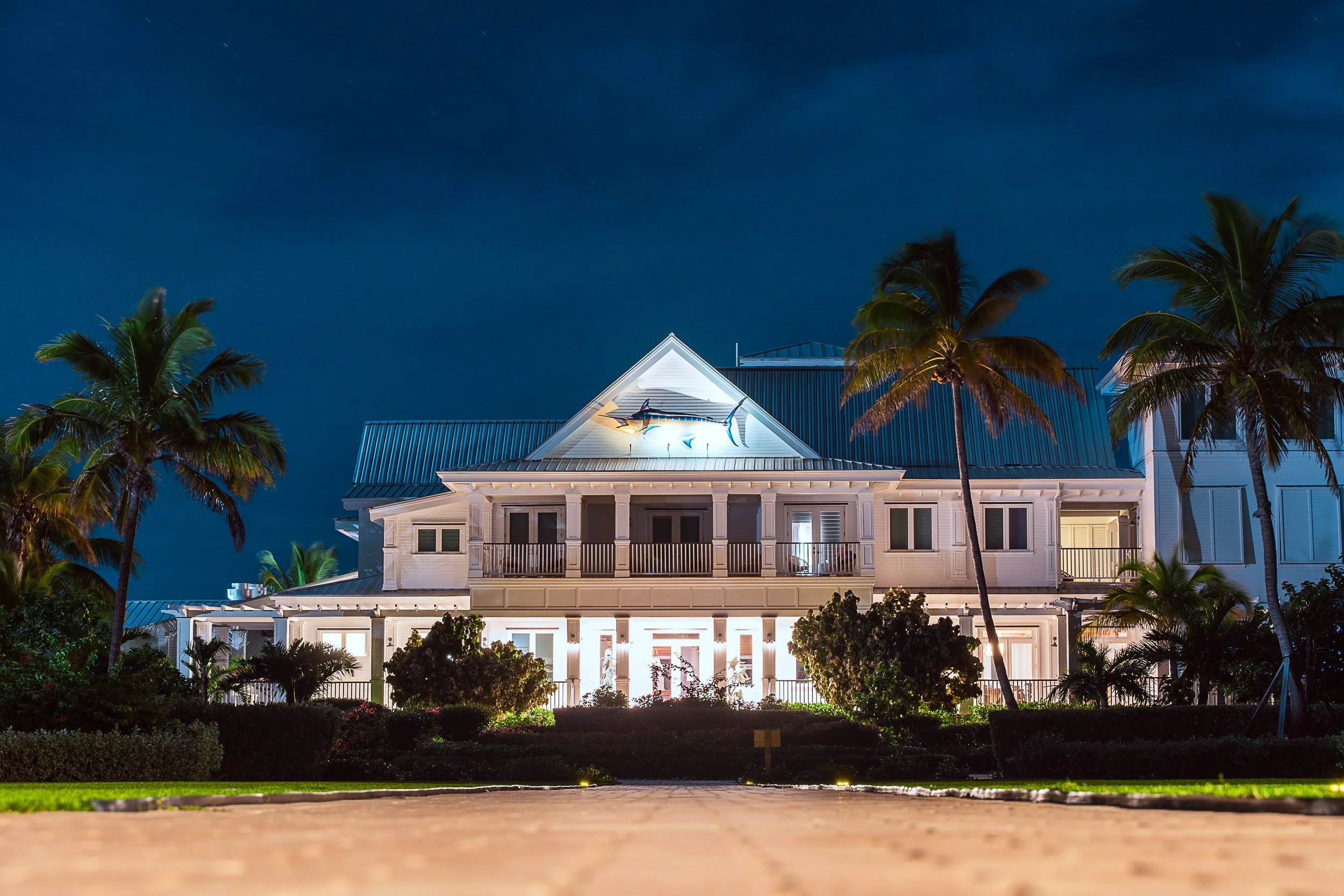 Large white house with illuminated exterior at night, surrounded by palm trees and landscaped gardens.