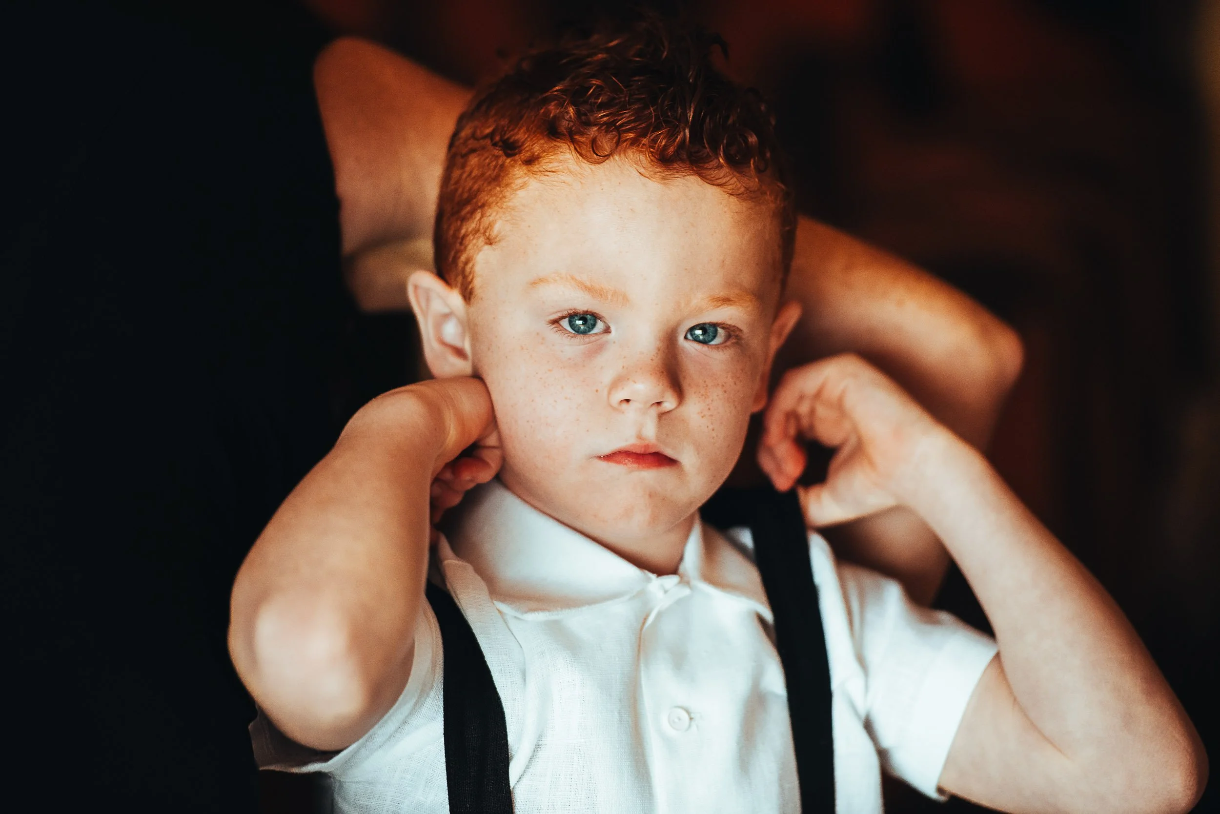 Boy adjusting suspenders, wearing a white shirt, with a serious expression.