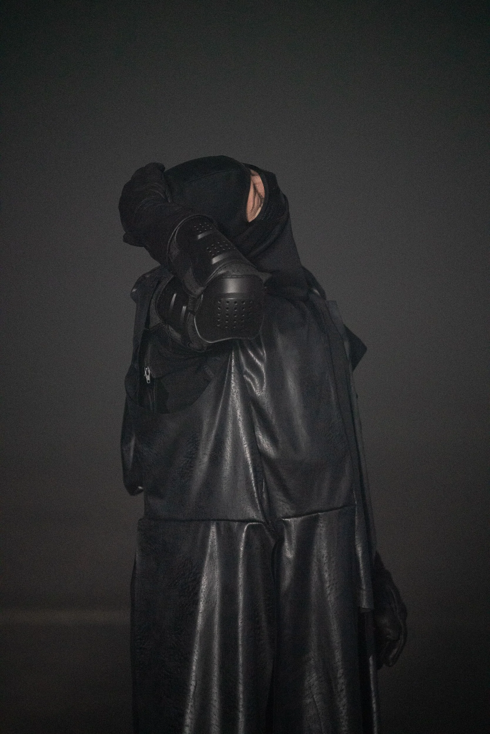 Person wearing black leather outfit and mask with hand covering part of their face against dark background.
