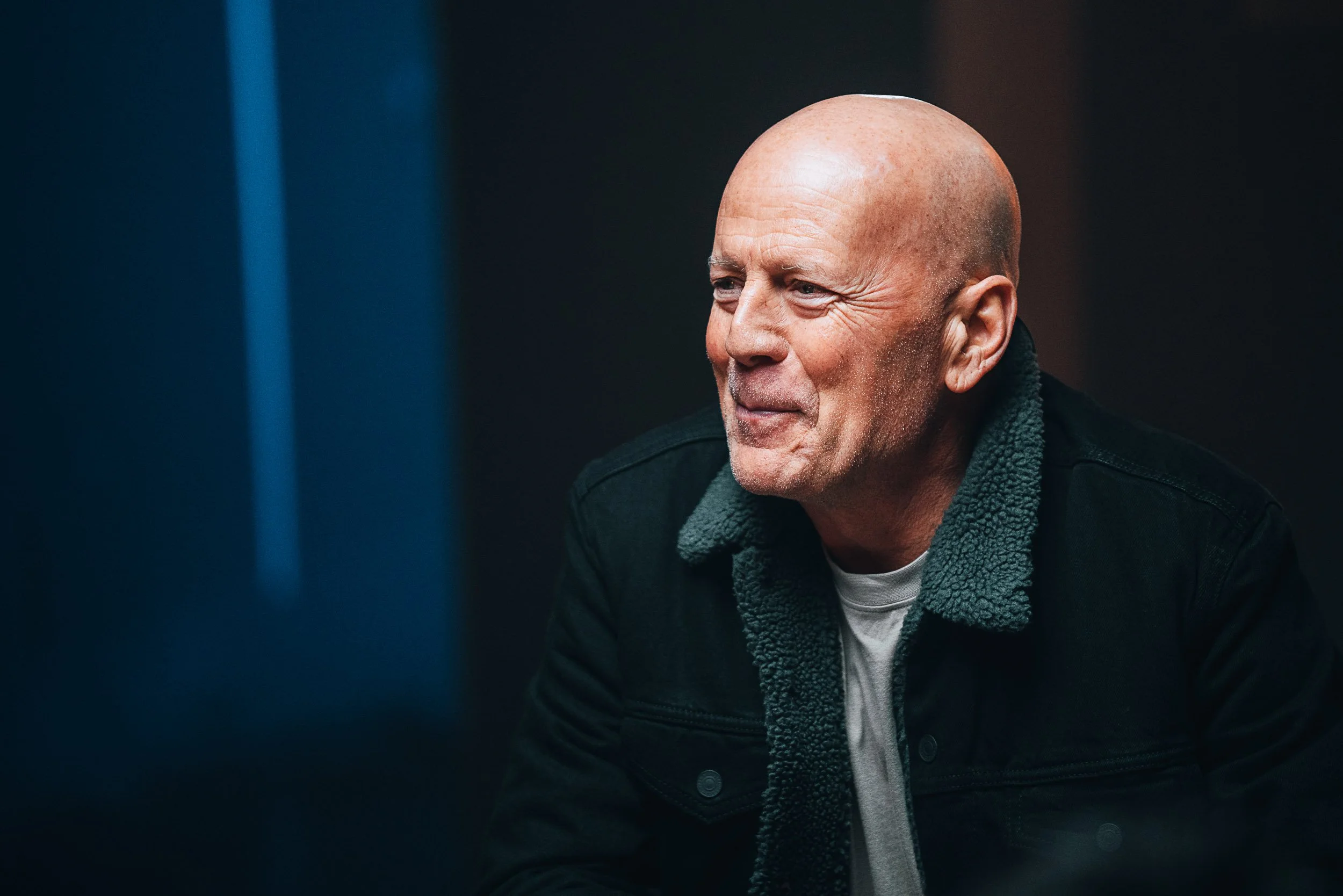 Person smiling with a shaved head wearing a black jacket with a fur-lined collar in a dimly lit setting.