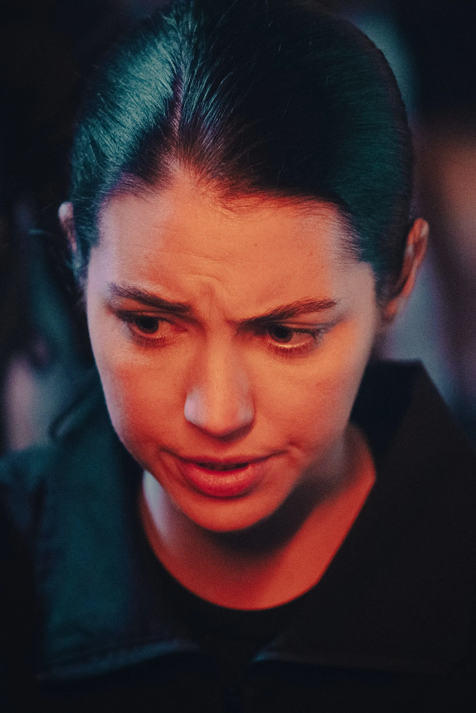 Close-up of a person looking downward with a focused expression, short dark hair, and wearing a dark top.