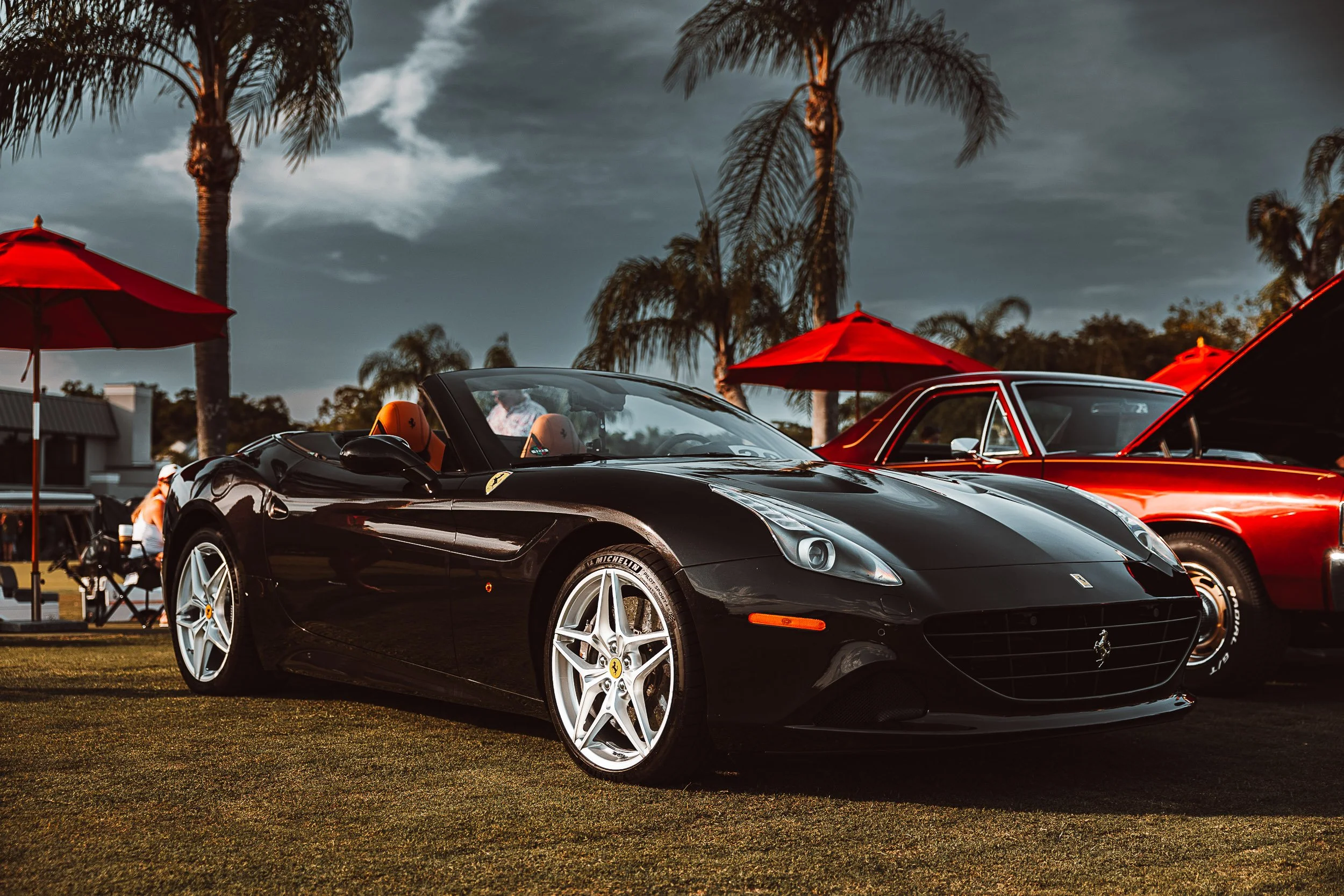 Black convertible sports car with red sports car in background, parked on grass, palm trees around, red umbrellas overhead, cloudy sky.