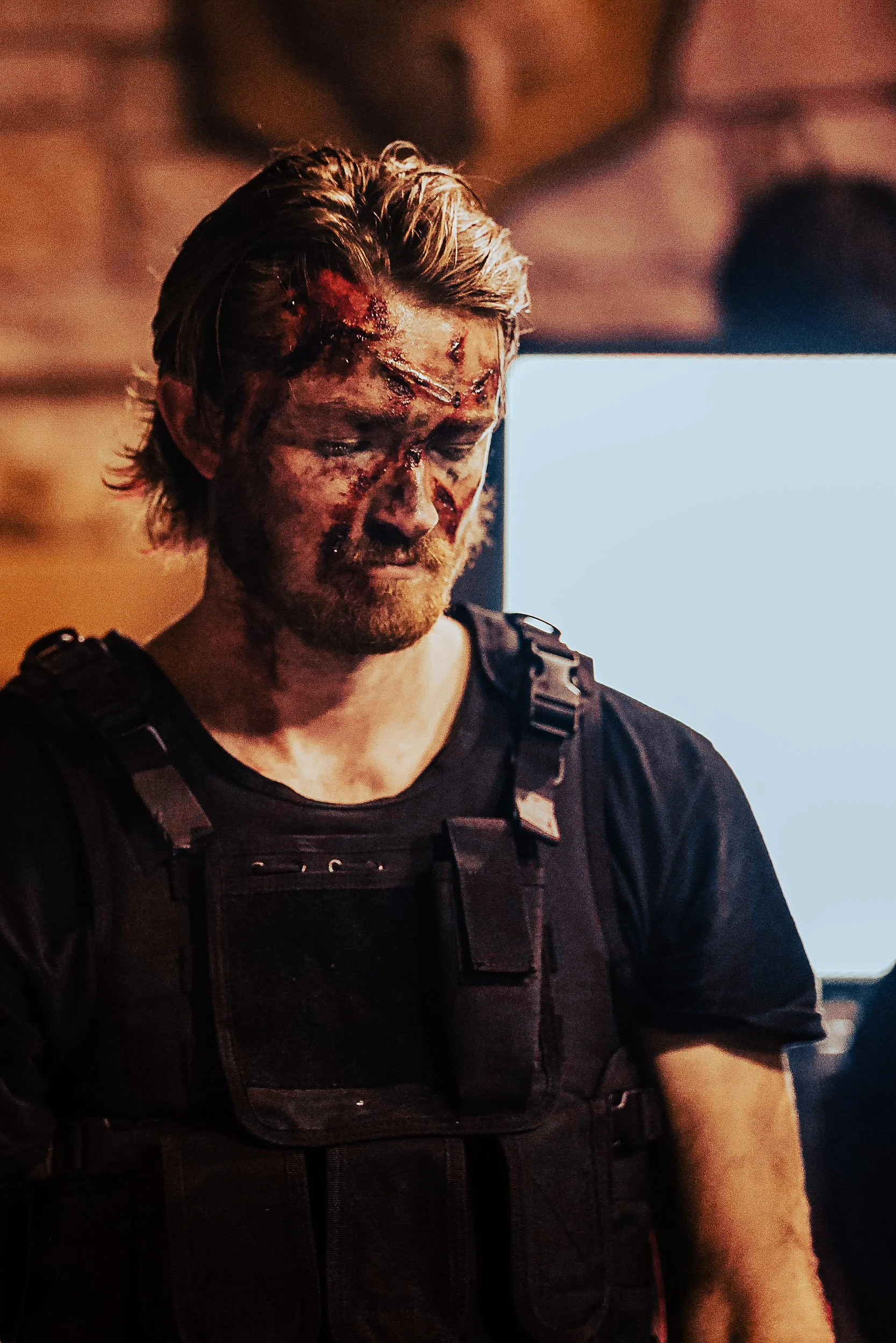 Person wearing tactical gear with blood and injuries on face, standing indoors.