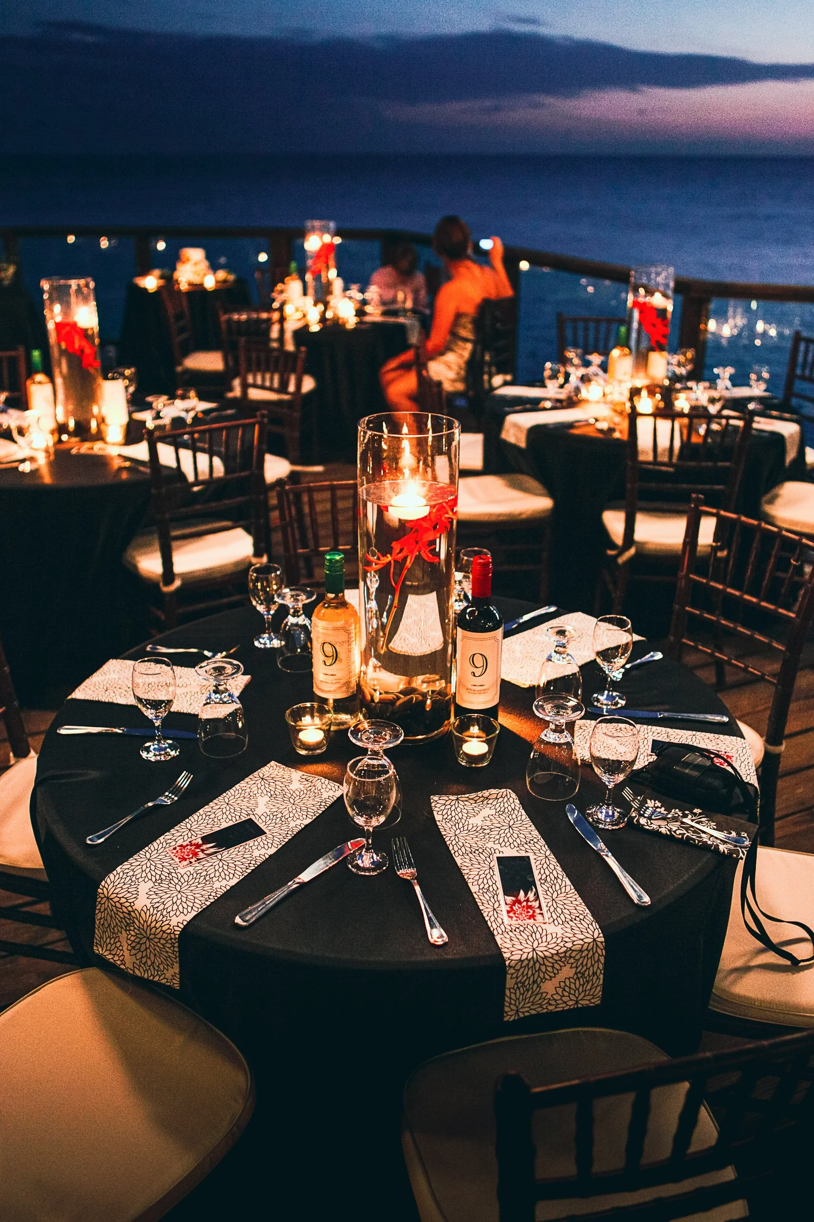Elegant outdoor dinner setting with round tables covered in black tablecloths decorated with candles, wine bottles, and place settings, set against a nighttime ocean backdrop.