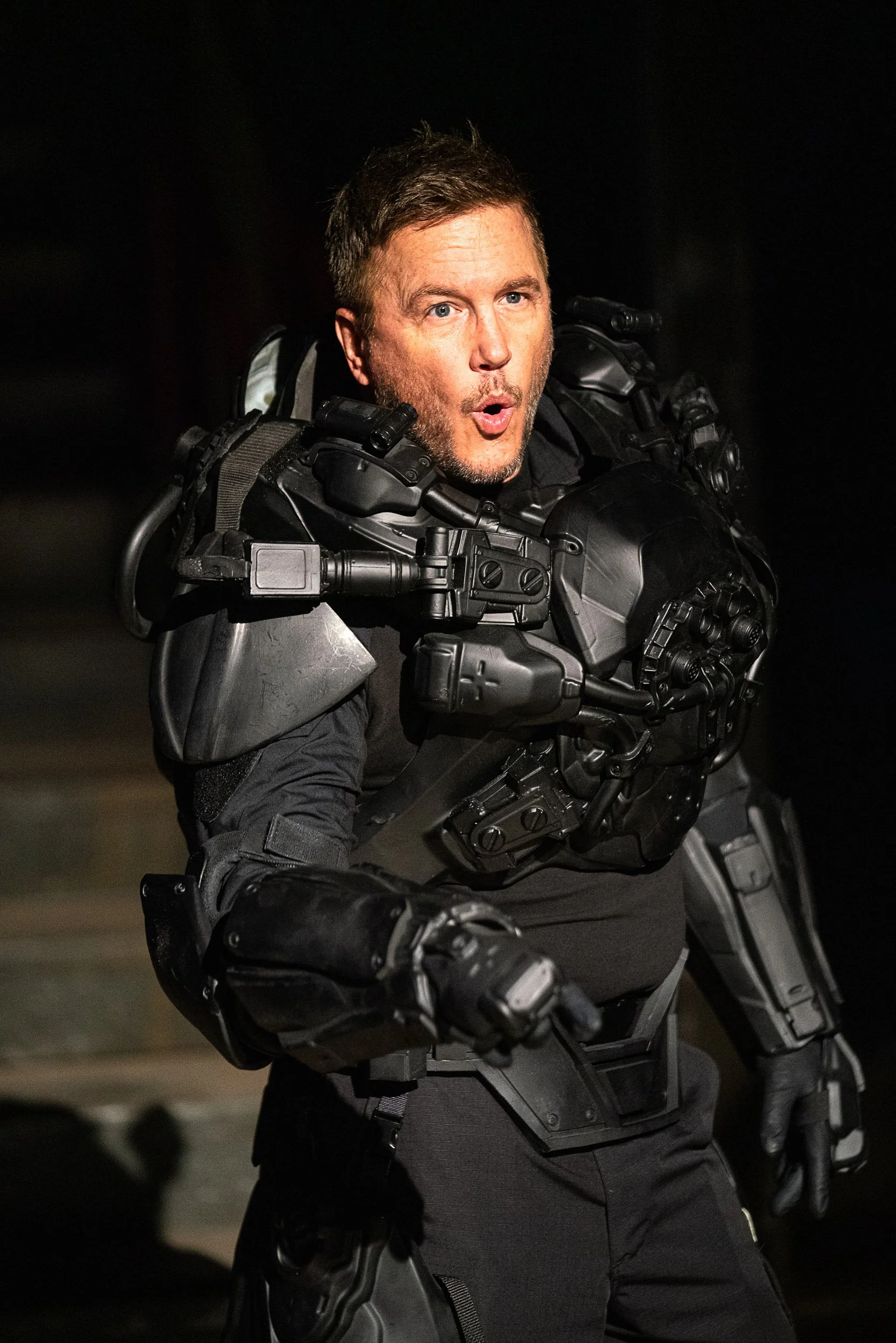 Person wearing a futuristic black armor suit, appearing surprised or shocked.