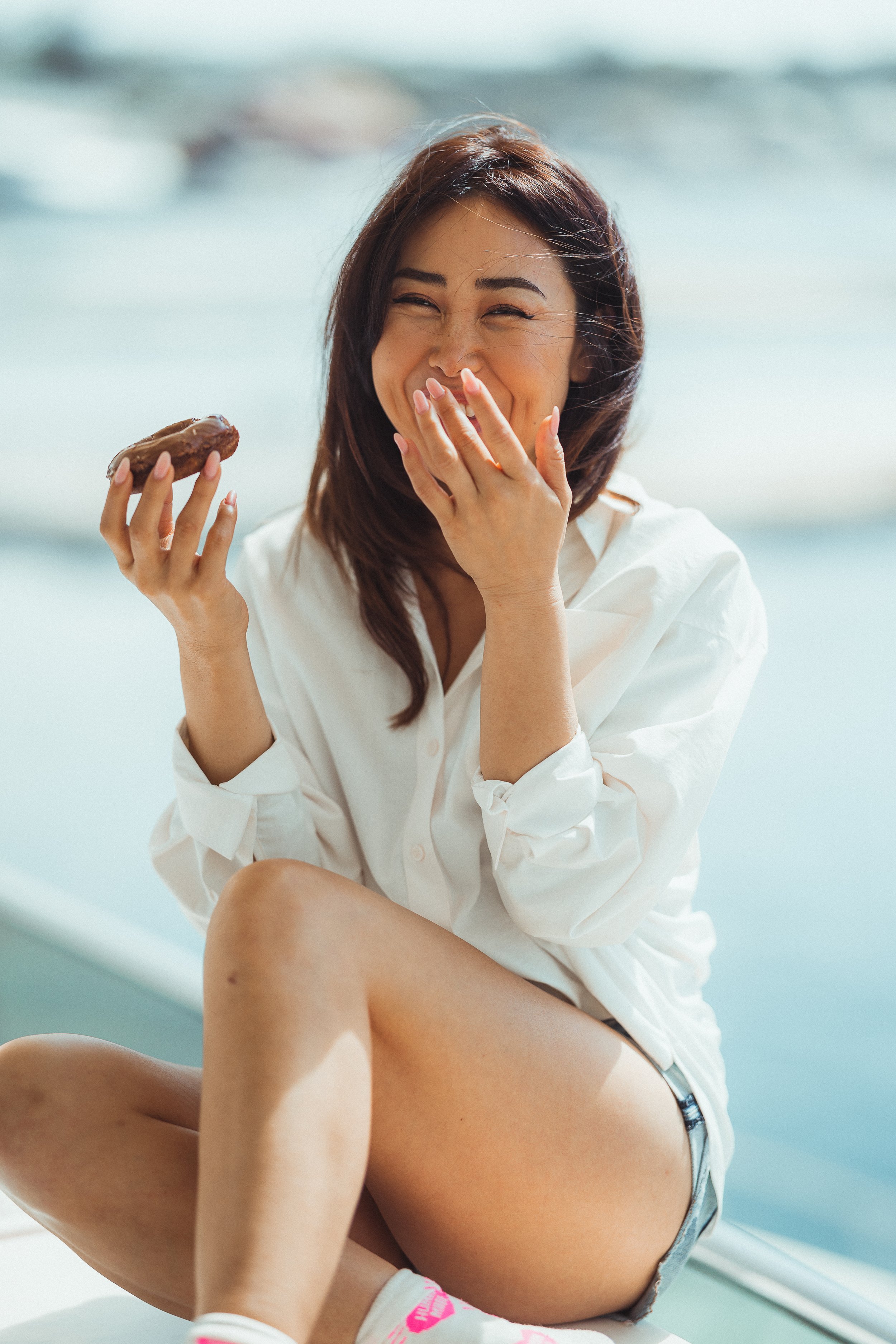 A woman with brown hair smiling and laughing, wearing a white shirt, sitting near water with waves in the background, holding a donut in her right hand, covering her mouth with her left hand.