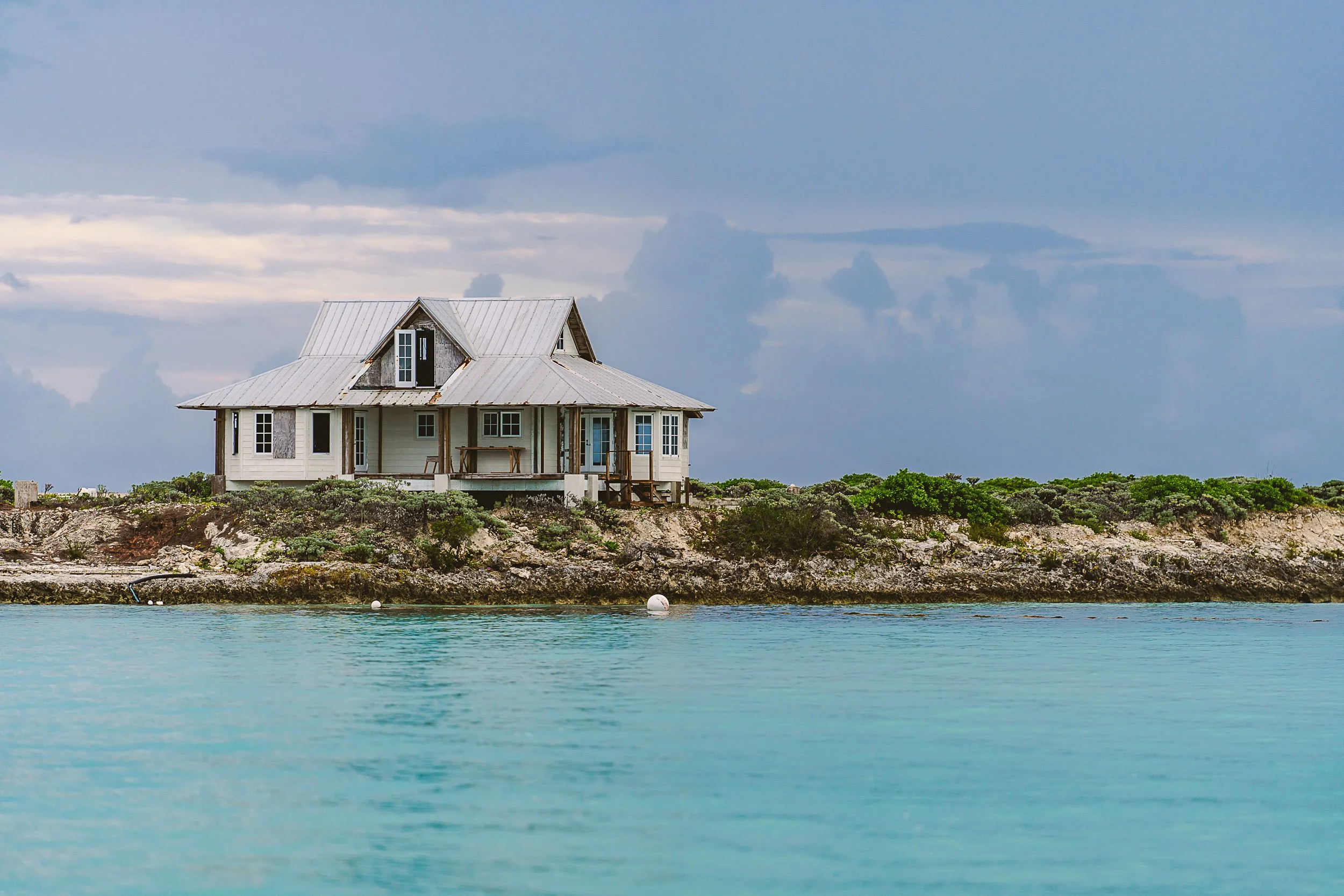 Wooden house on a rocky coastal shore with a metal roof, surrounded by greenery and turquoise water in the foreground.
