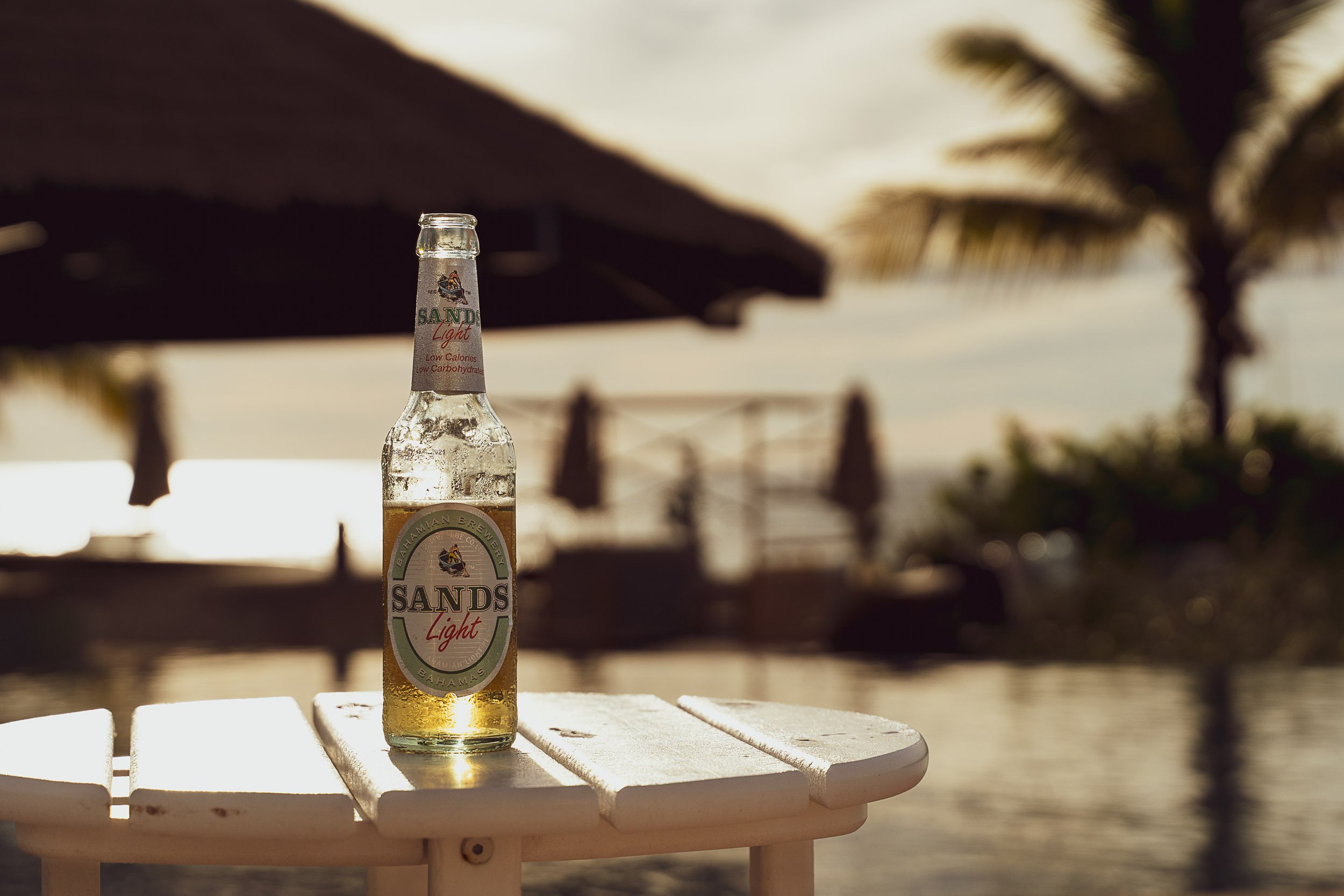 Sands Light beer bottle on outdoor table with tropical background, palm tree, and thatched roof.