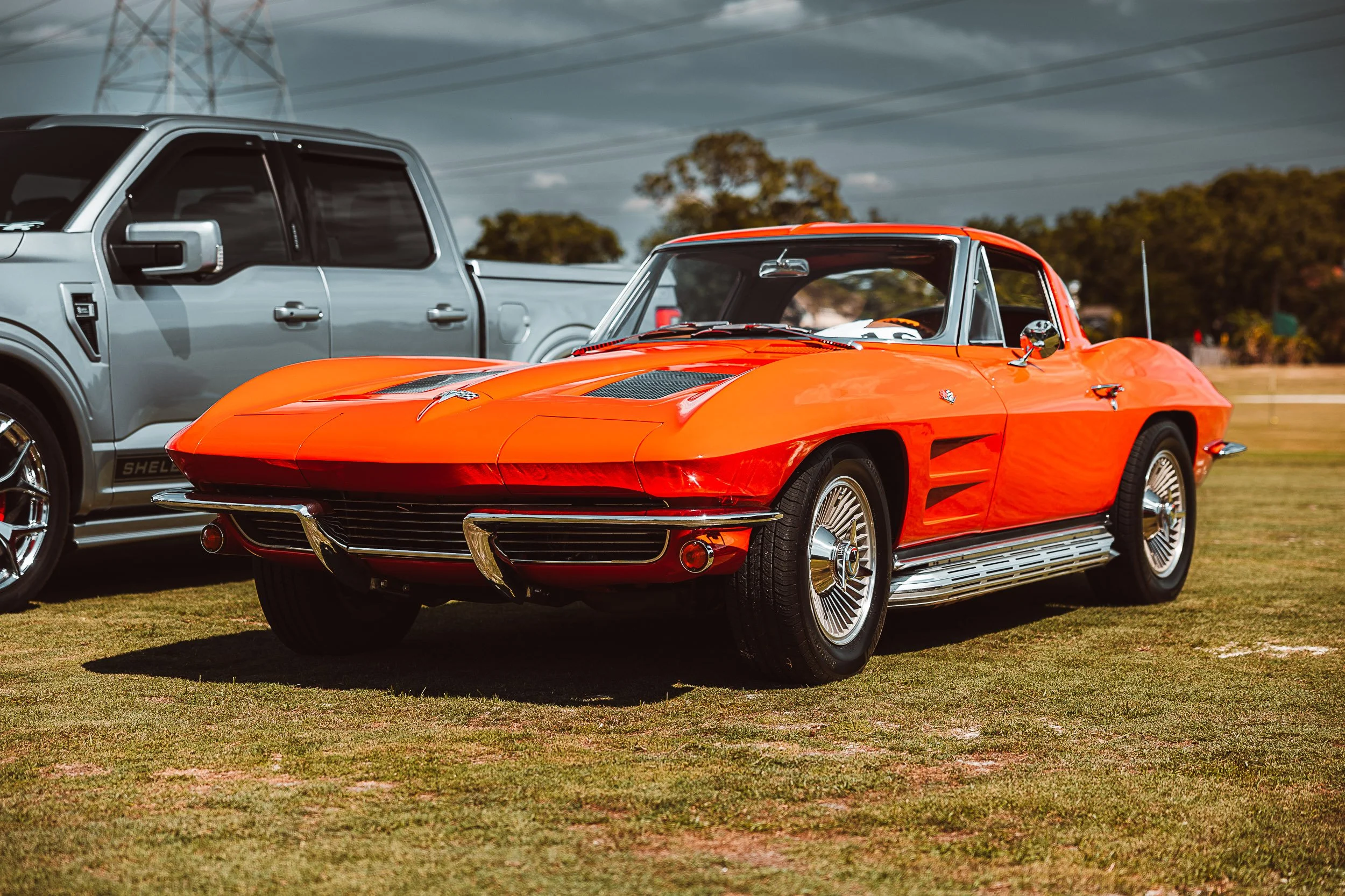 A bright orange vintage sports car parked on grass next to a silver pickup truck under a cloudy sky.