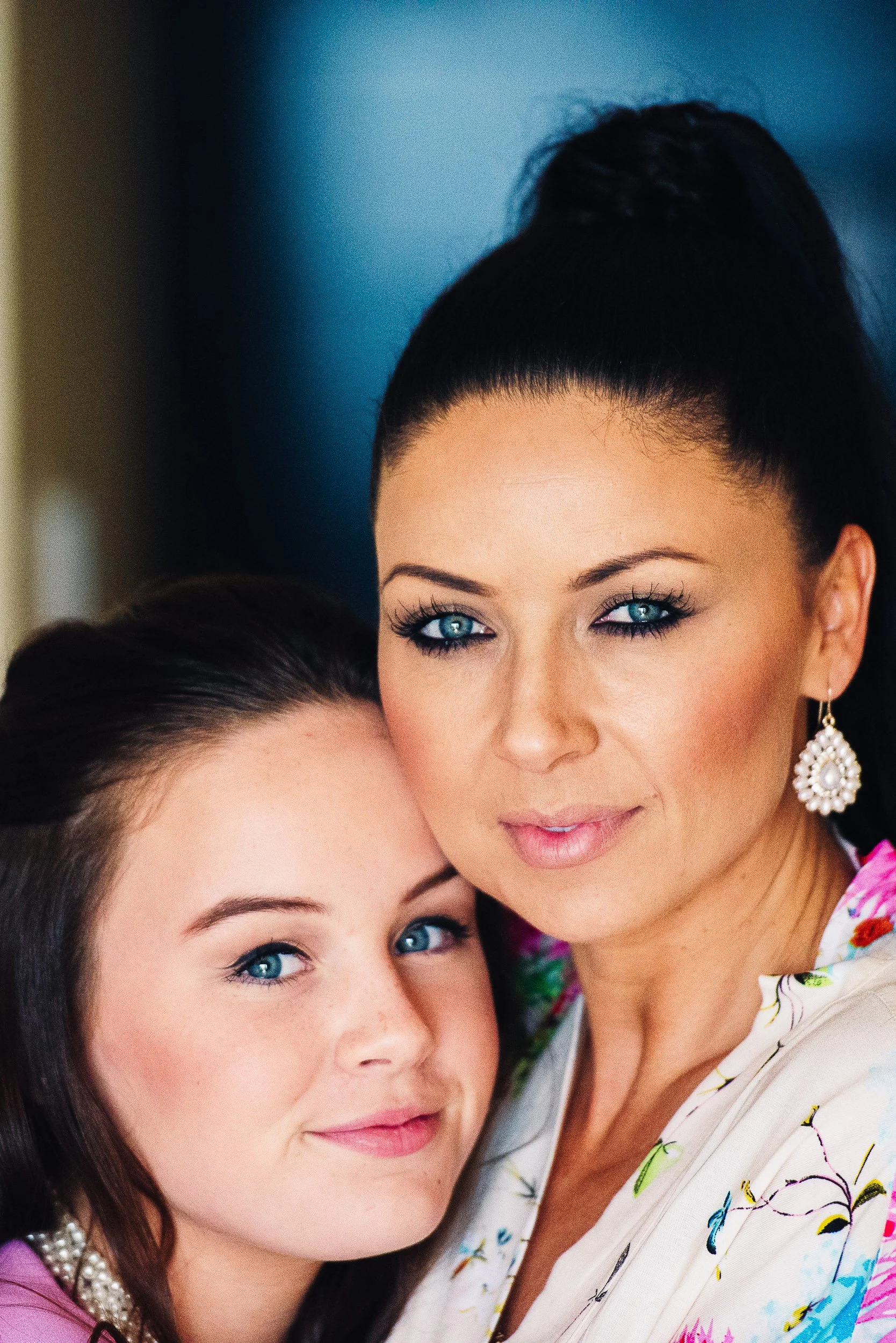 A woman with dark hair and blue eyes wearing earrings, next to a young girl with similar features, both smiling slightly.