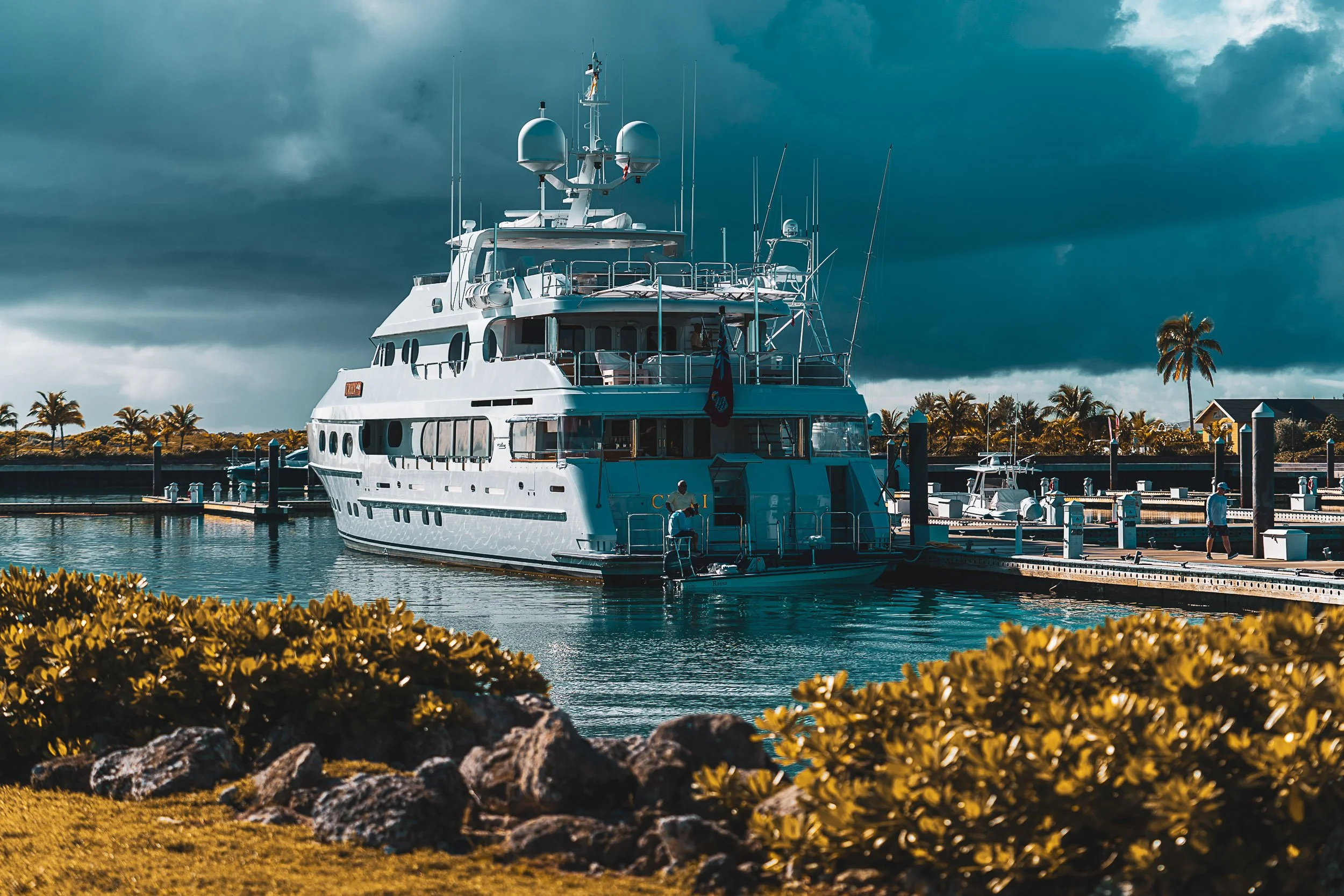 Luxury yacht docked in marina with people and lush greenery, under cloudy sky.