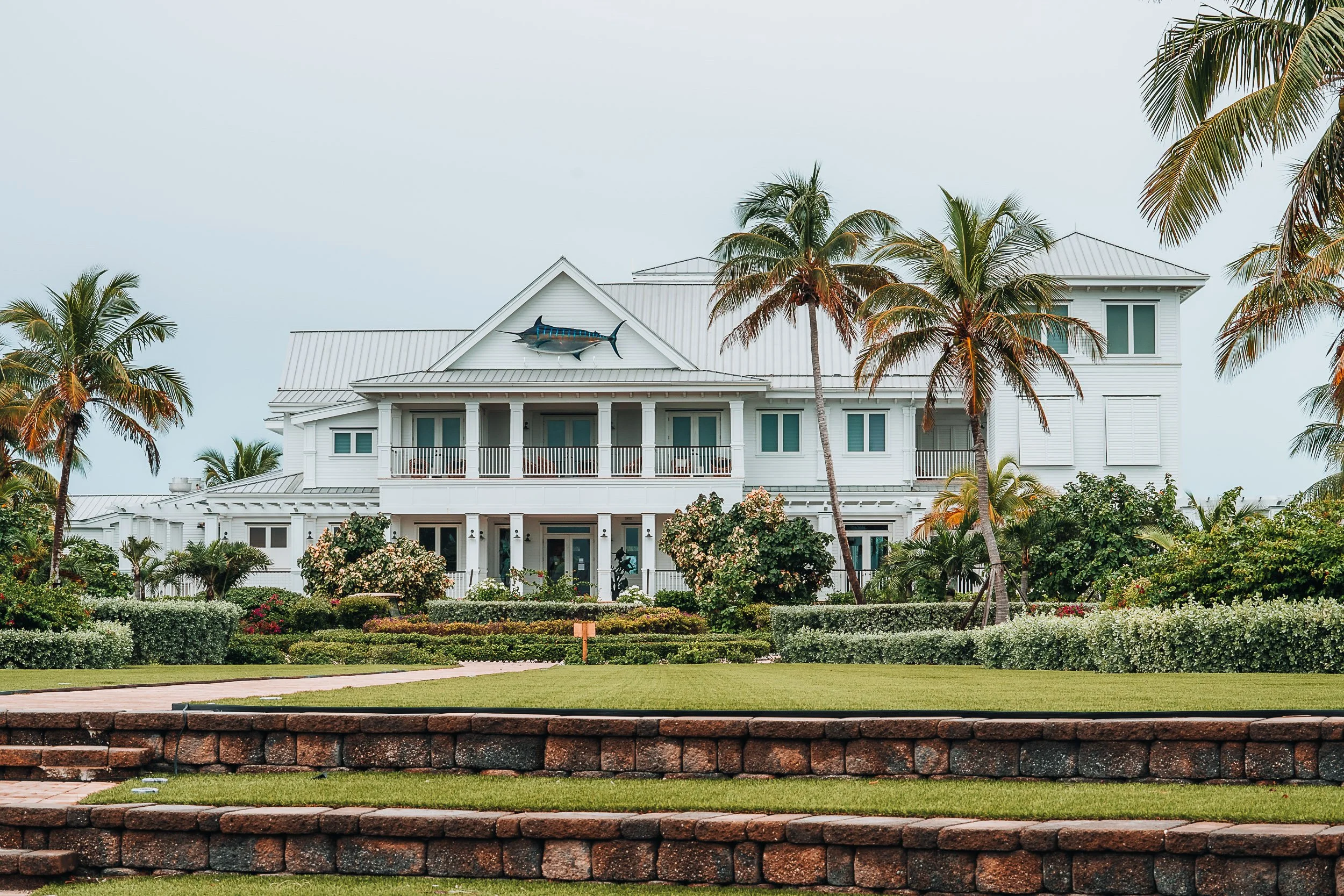 Large white house with a porch, surrounded by palm trees and landscaped gardens, featuring a decorative fish on the front gable.