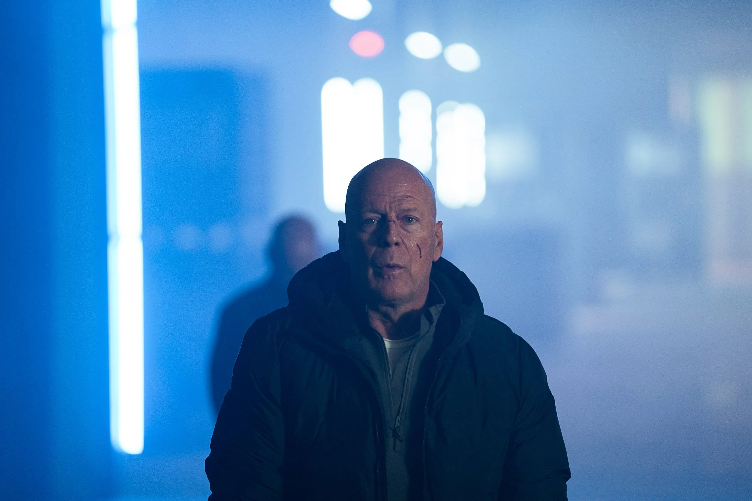 Bald man in jacket standing in a dim, blue-lit indoor setting with blurred lights in the background.
