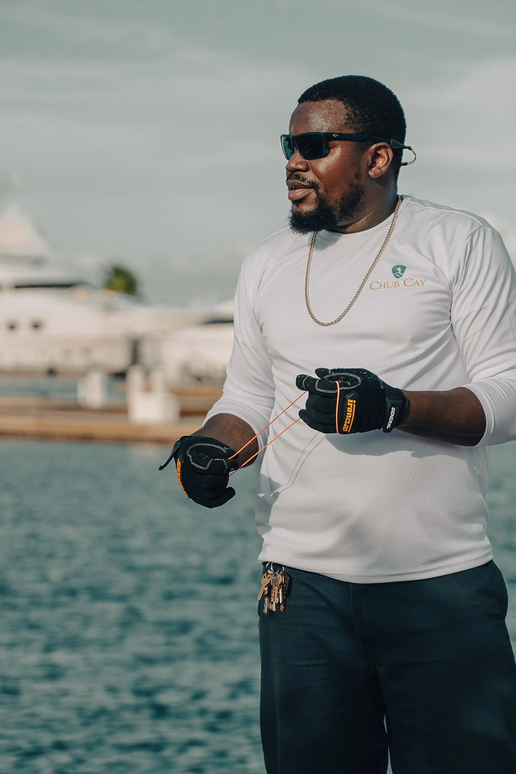 Man in sunglasses and gloves near water with boats in the background.