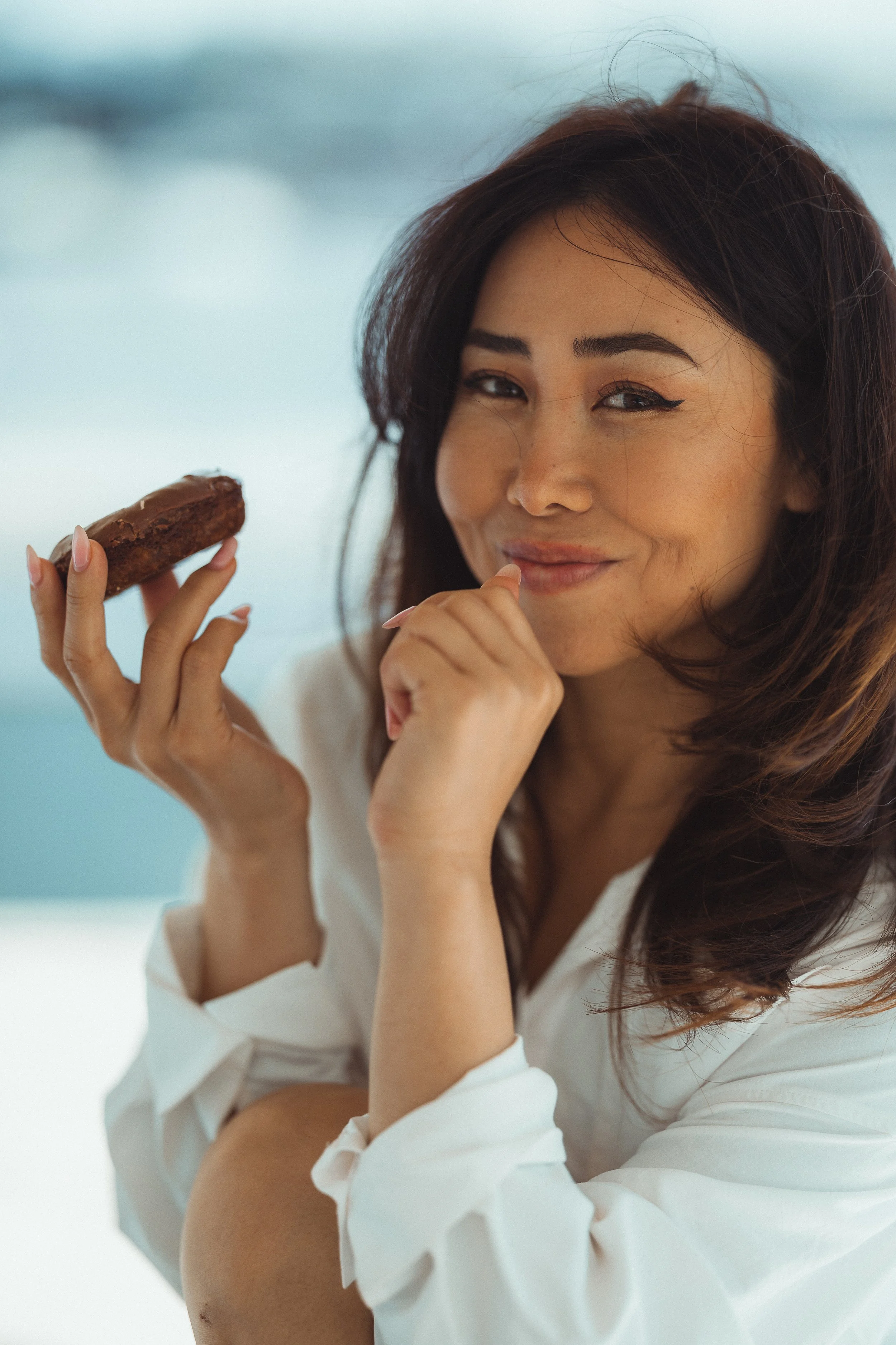 A woman with dark wavy hair and winged eyeliner holds a chocolate-covered ice cream bar and smiles at the camera while resting her chin on her hand, wearing a white shirt, against a blurred light background.