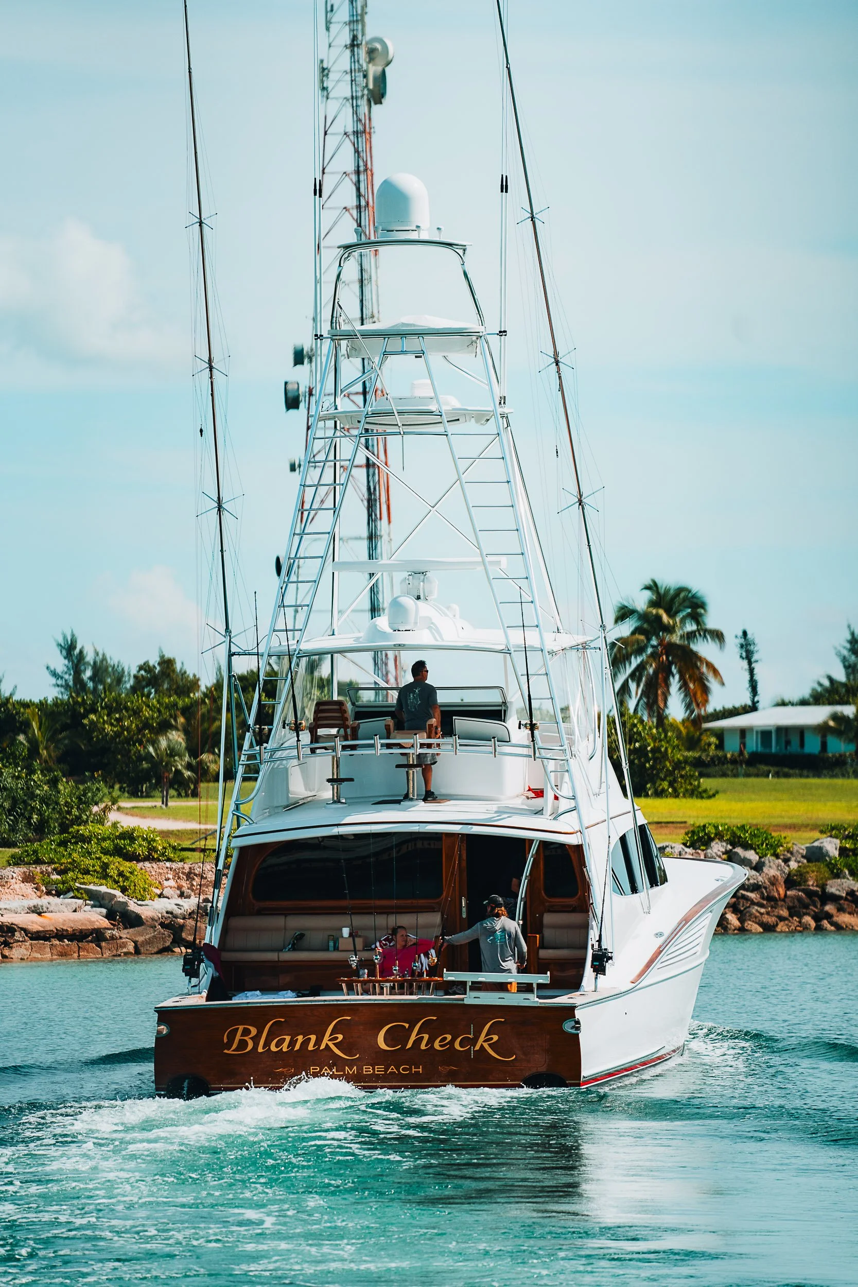 Luxury yacht "Blank Check" cruising on a waterway, with people on deck, green landscape and palm trees in the background.