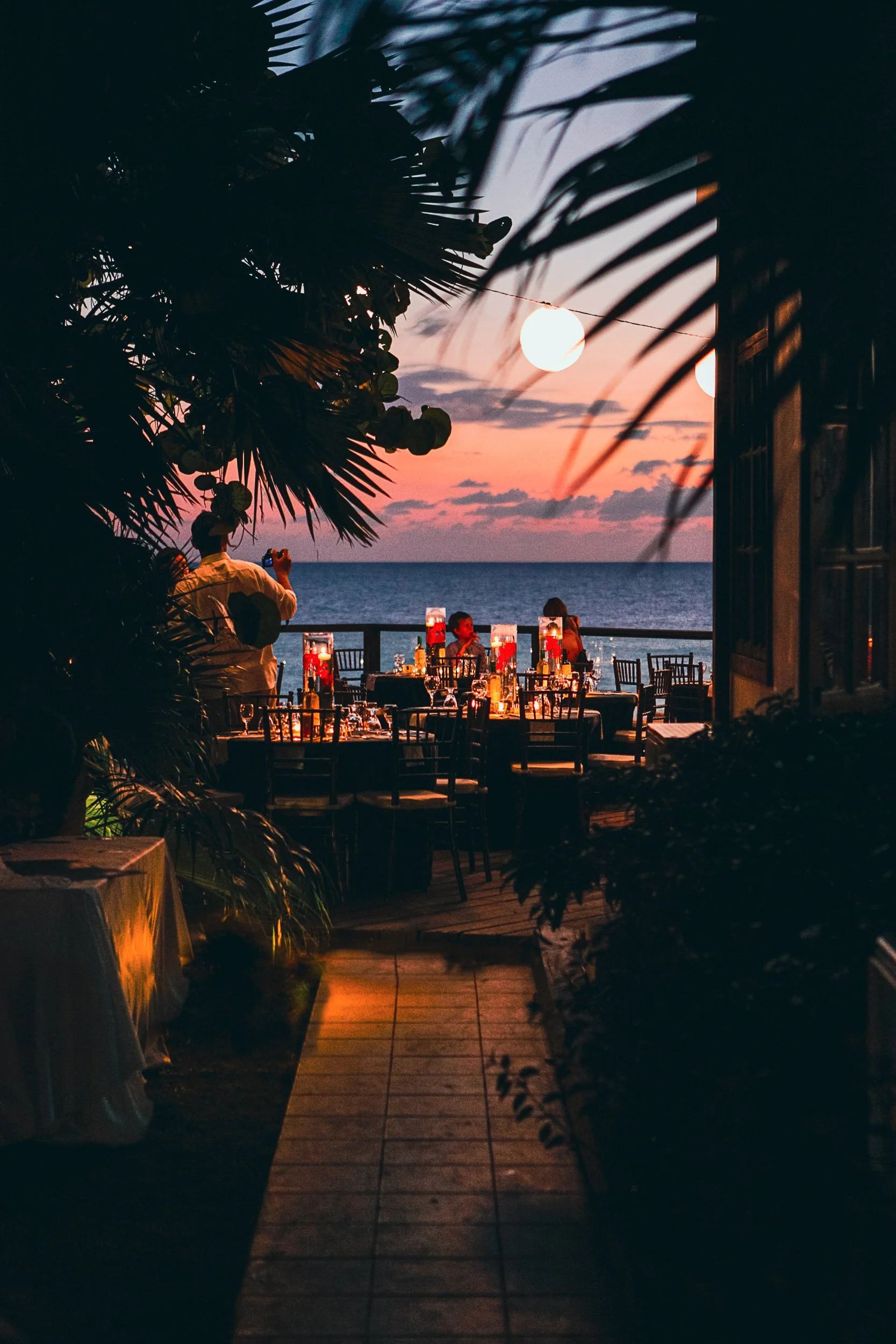 Outdoor dining scene at sunset with tables set for dinner, palm trees, string lights, and an ocean view.