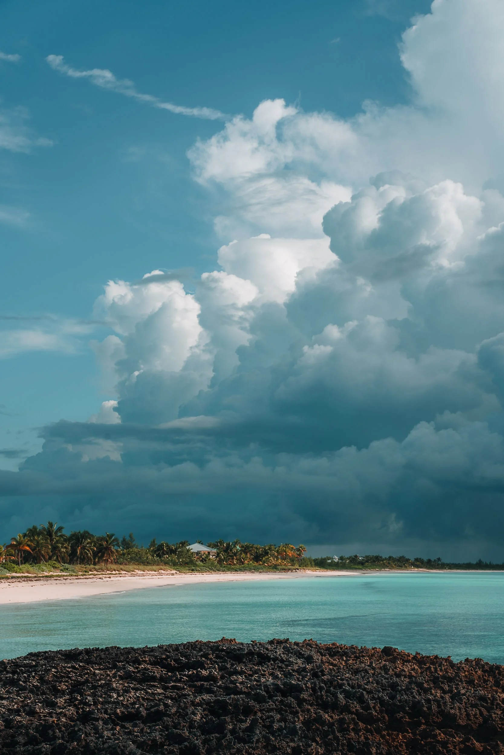 Tropical beach with turquoise water, a rocky shoreline in the foreground, palm trees in the distance, and large cumulus clouds in the sky.
