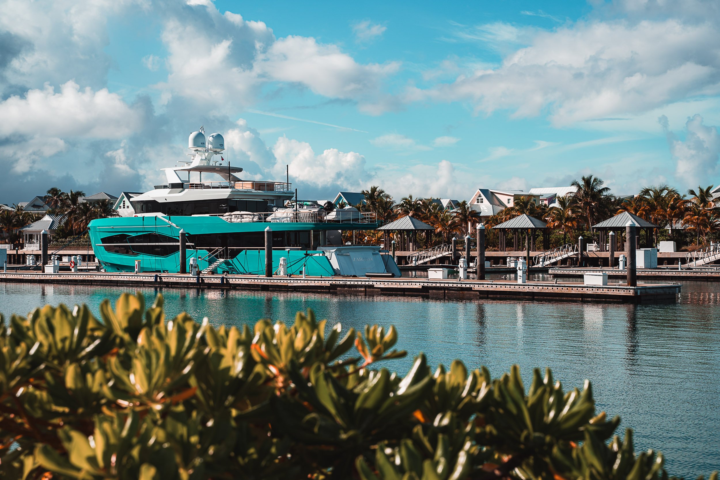 A large luxury yacht docked at a marina, with palm trees and houses in the background, under a bright blue sky with clouds.