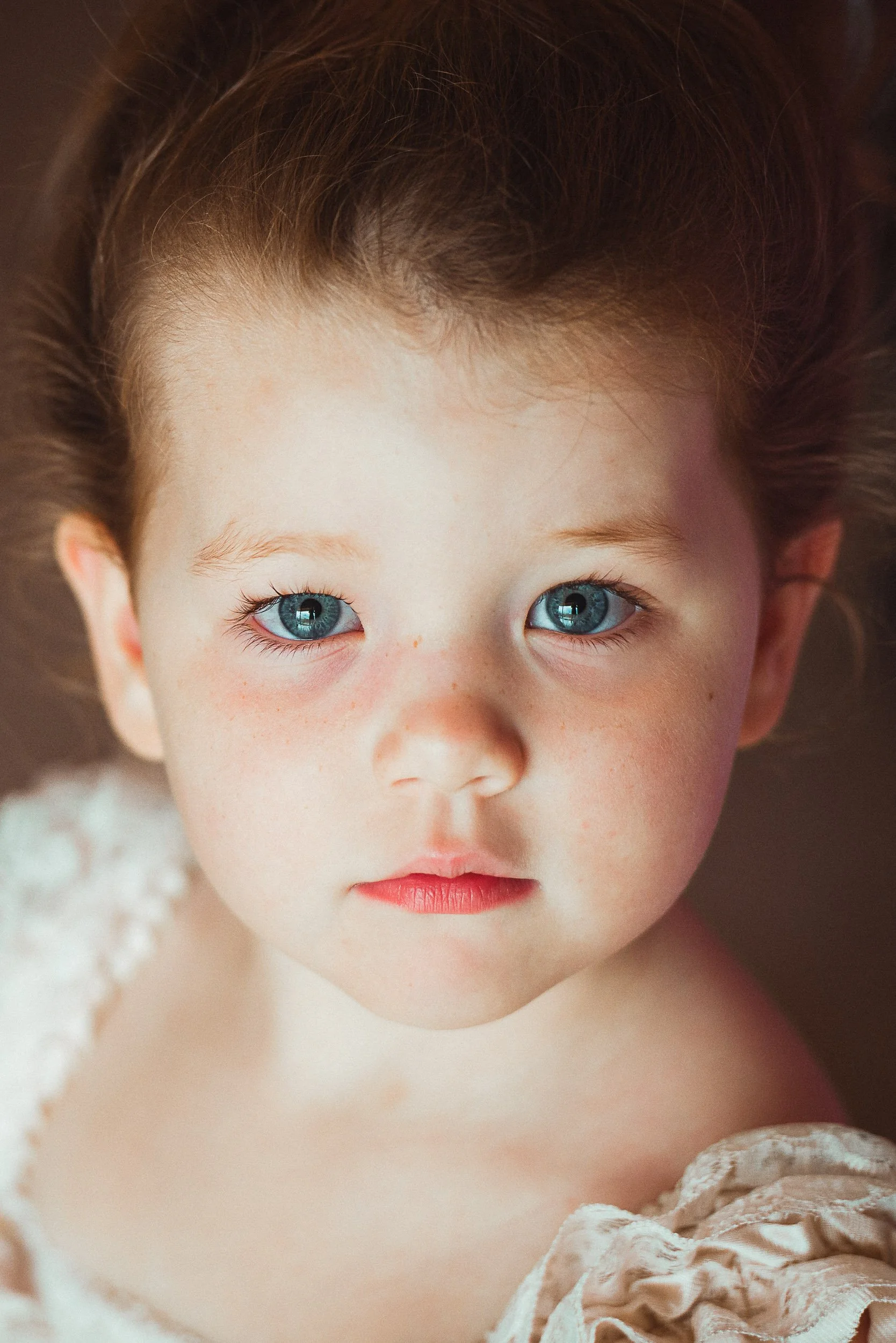 Close-up of a young child's face with blue eyes, fair skin, and light brown hair, wearing a white garment.