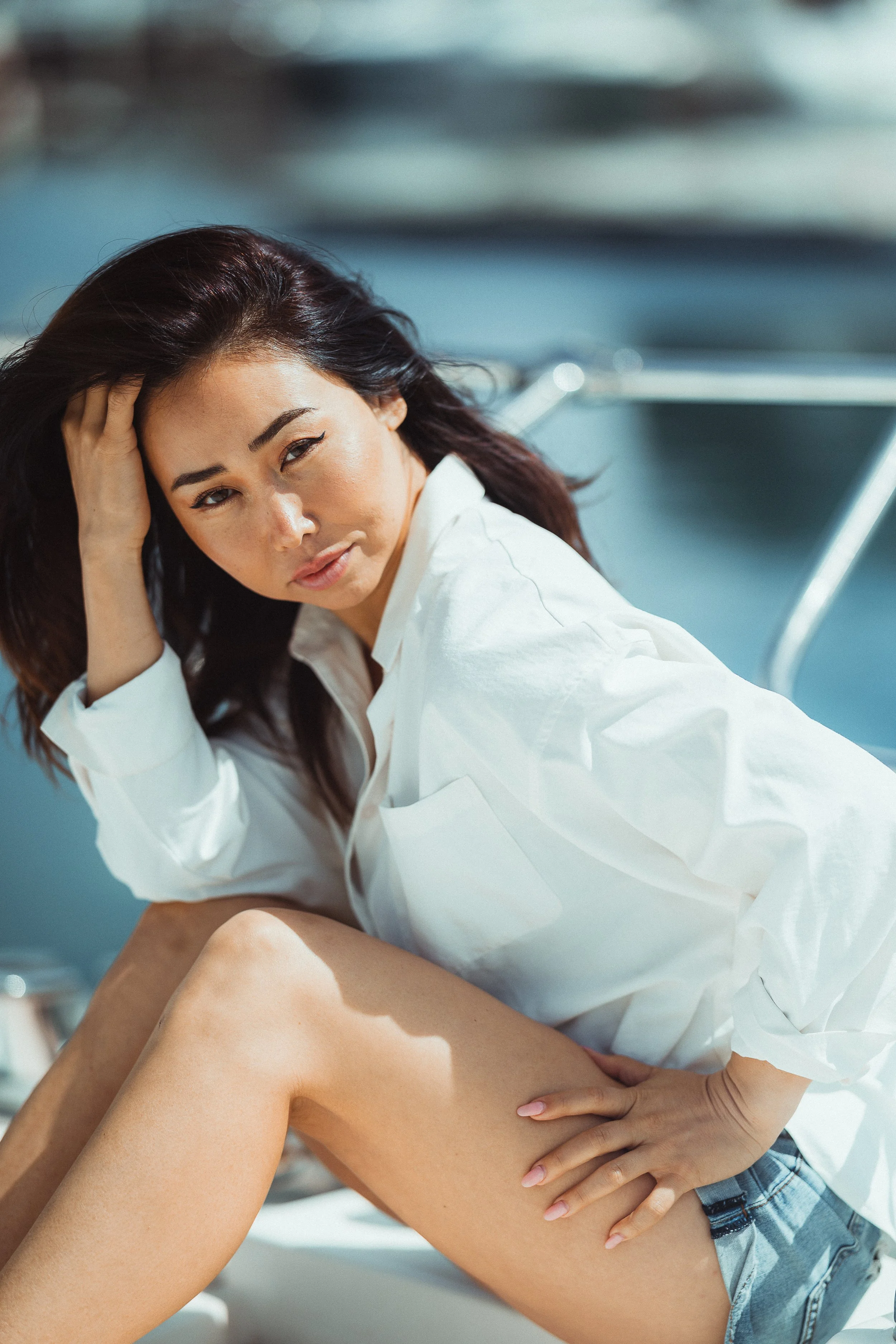 A woman with dark hair and light skin, wearing a white blouse and denim shorts, sitting outdoors near a body of water, with sunlight highlighting her face and legs.