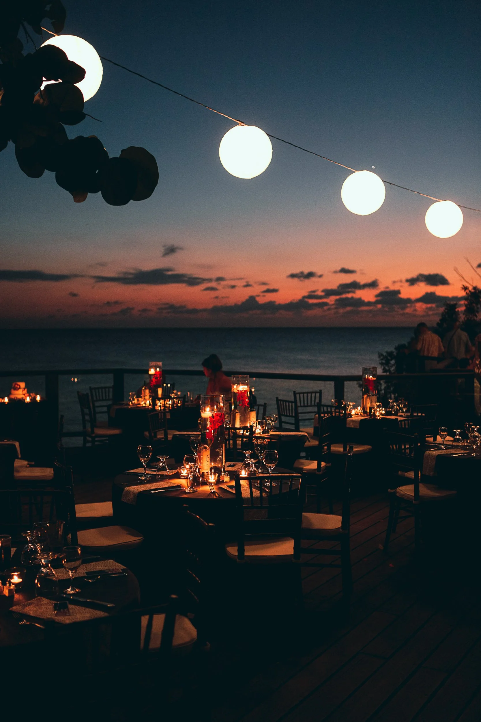 Outdoor beachside dining area at sunset with round tables, chairs, and string lights. Glasses and candles are set on tables, creating a cozy, romantic atmosphere.