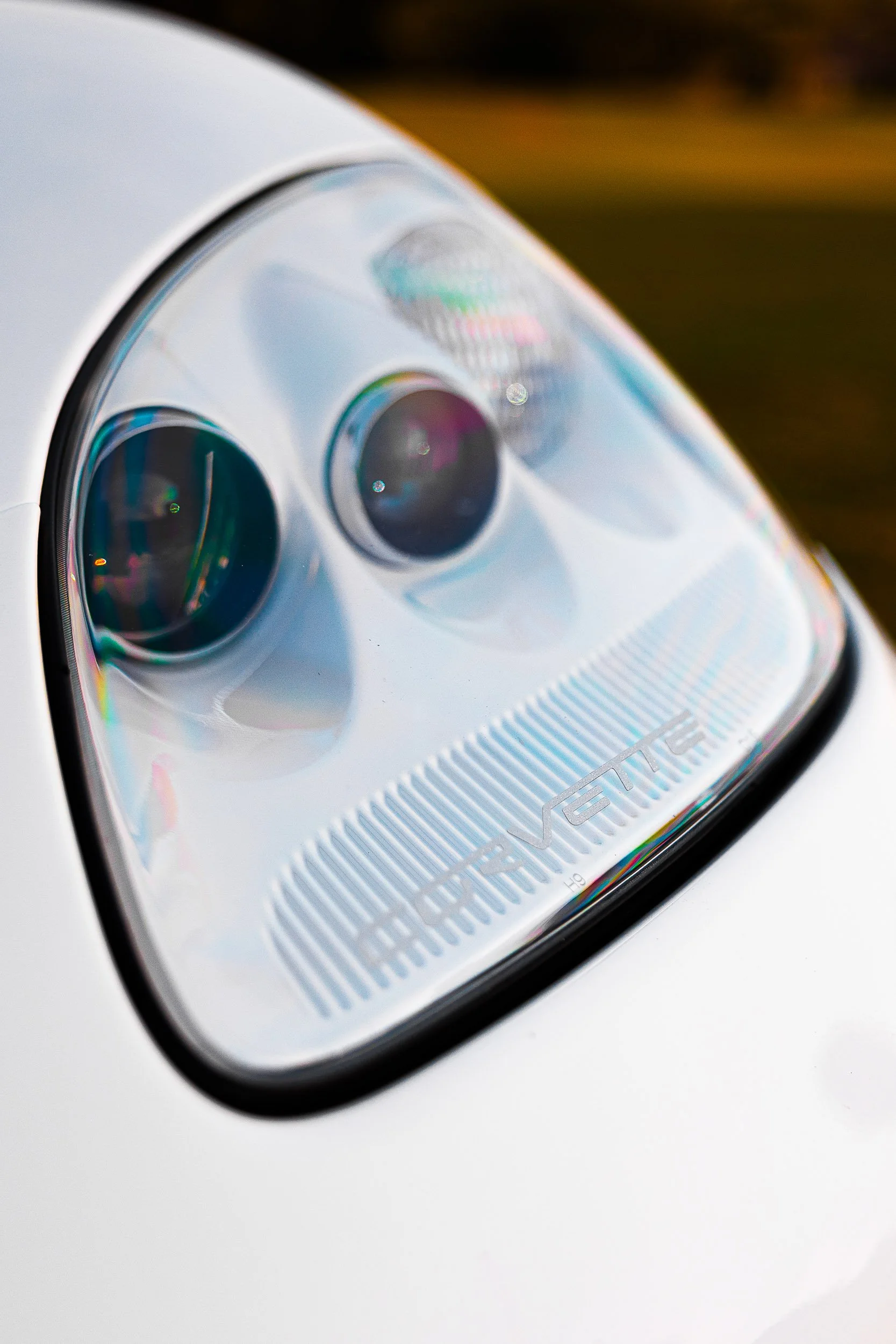 Close-up of a car headlight with "Corvette" branding visible on the lens, against a blurred background.