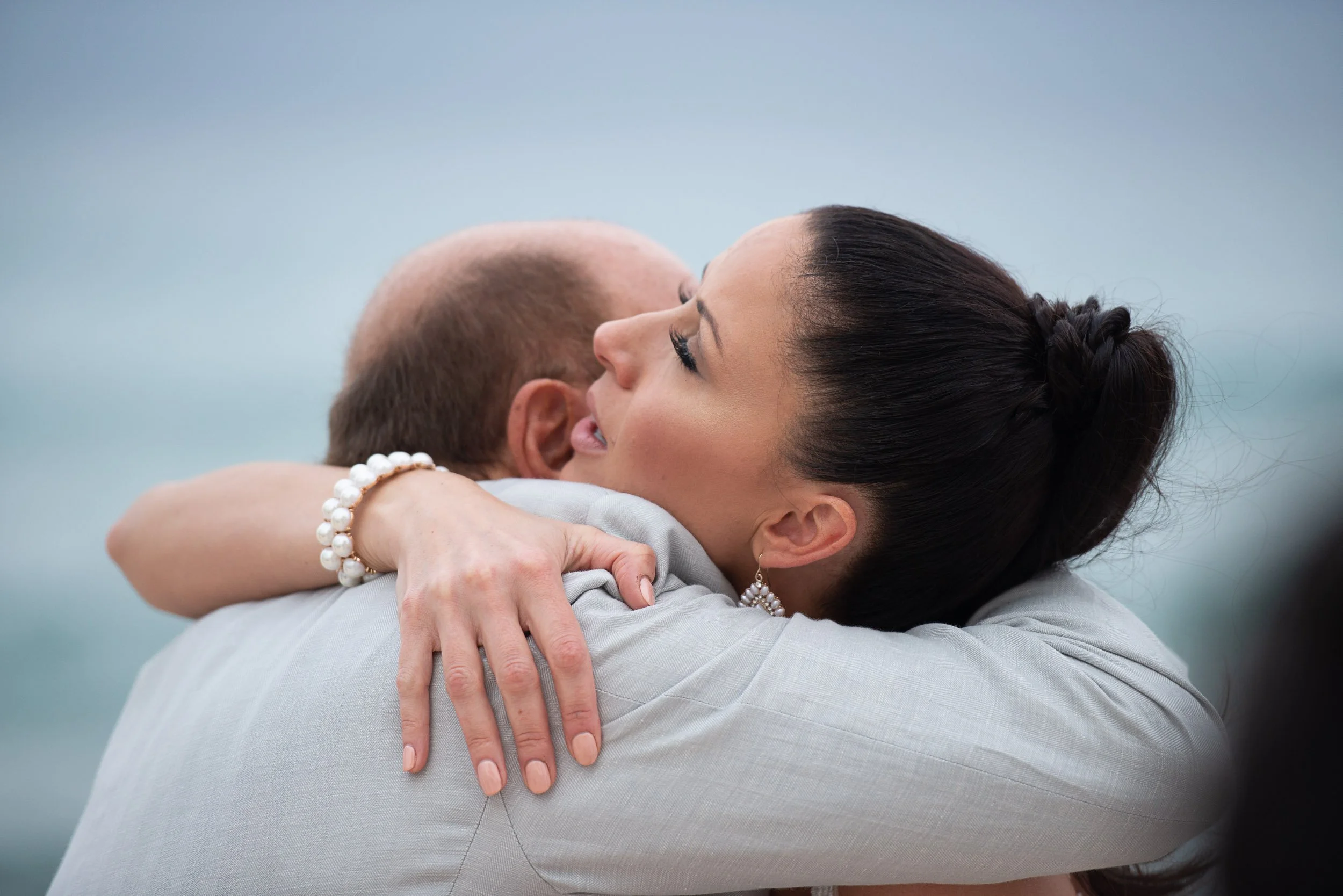 Woman and man hugging closely, woman wearing pearl bracelet and earrings, man's back turned.