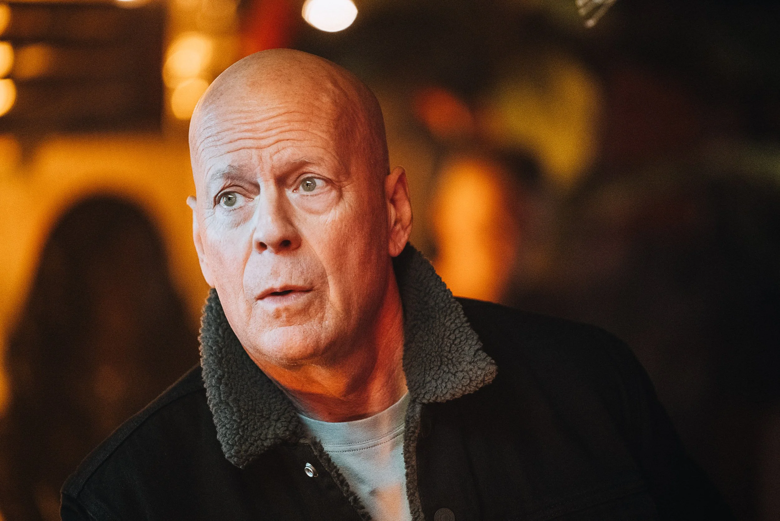 Bald man in a jacket with a fleece collar, indoors with blurred warm lighting in the background.