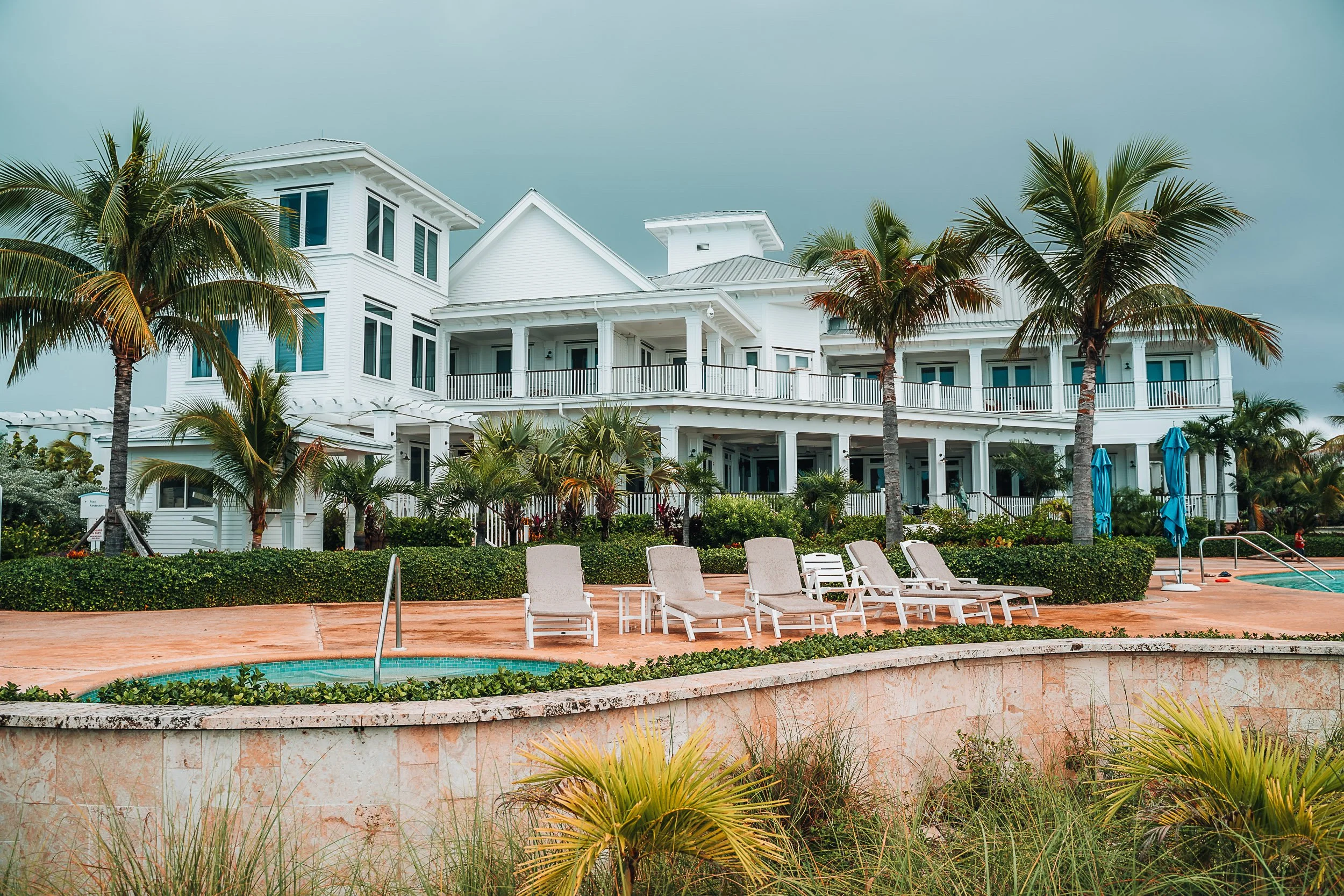 Luxurious white beach house with palm trees, pool, and lounge chairs in a tropical setting.