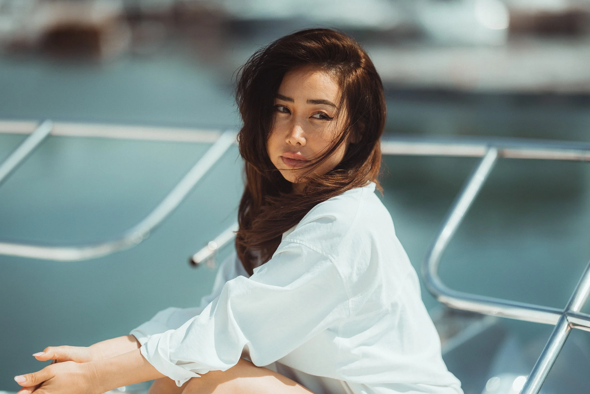 A woman with brown hair and light skin sitting outdoors by a marina, with blurred boats in the background, wearing a white long-sleeve shirt and looking to her left.