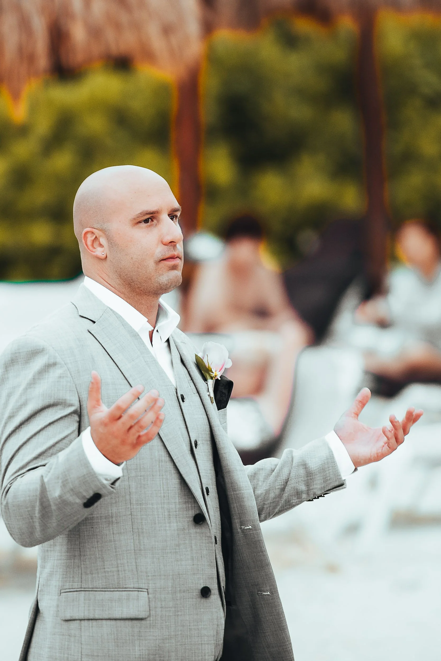 Man in gray suit with floral boutonniere, standing outdoors, gesturing with open hands.