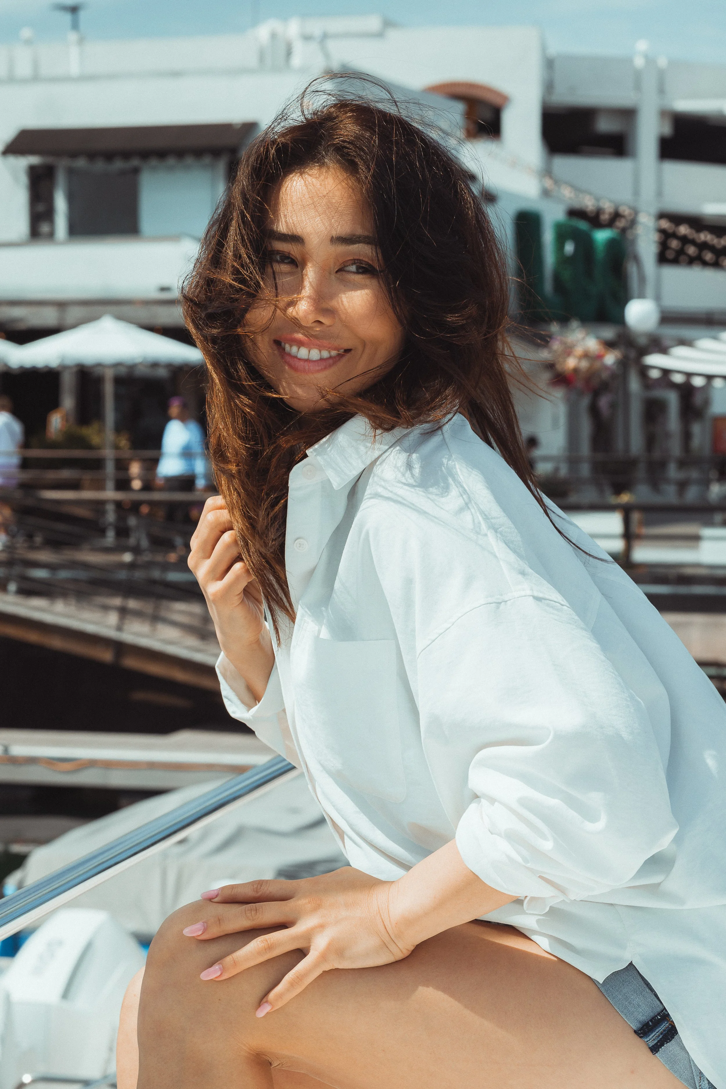 A woman with brown hair and a white shirt sitting outdoors, receiving a massage or treatment on her shoulder and back.
