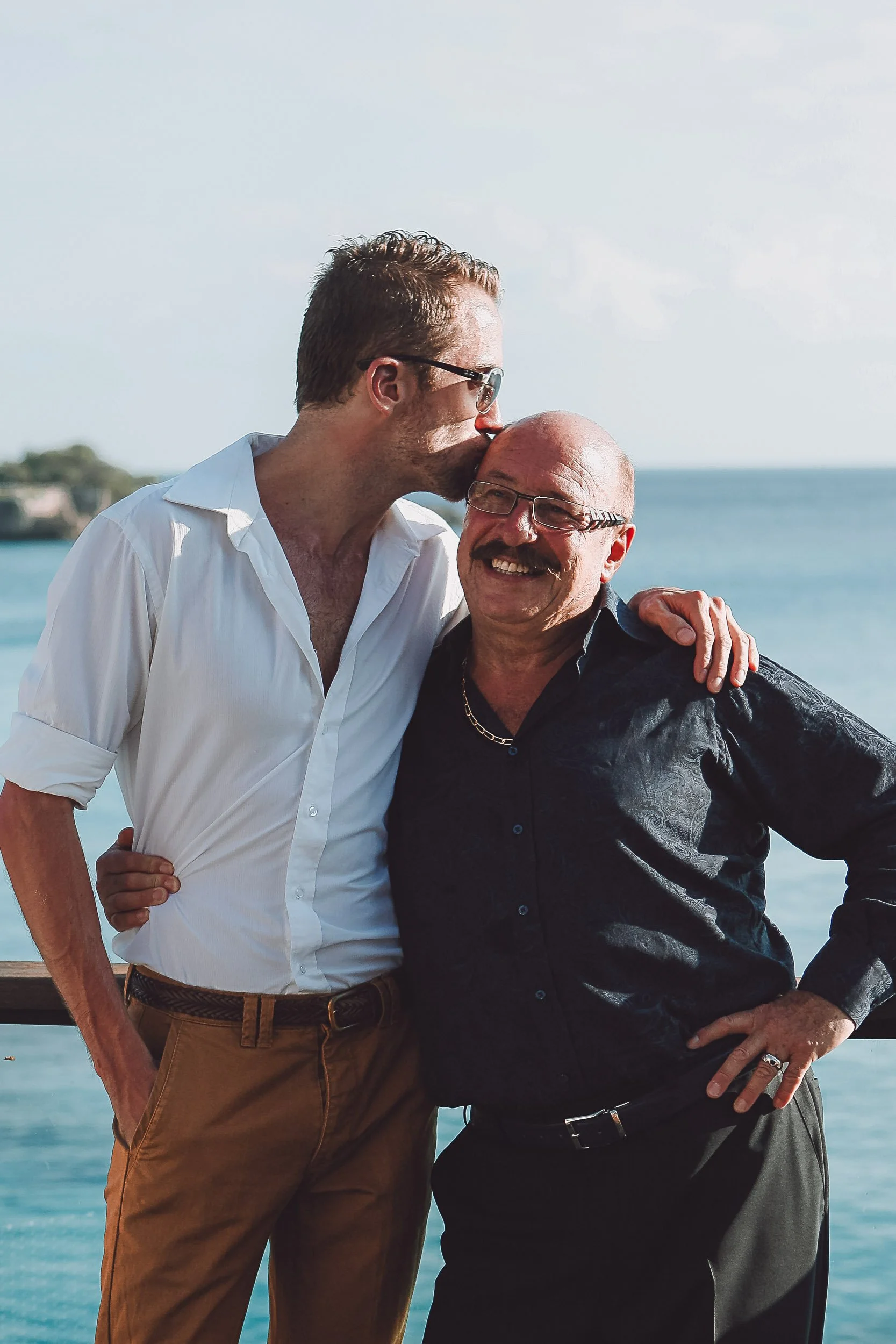 Two men smiling and embracing near the ocean, with one kissing the other's head.